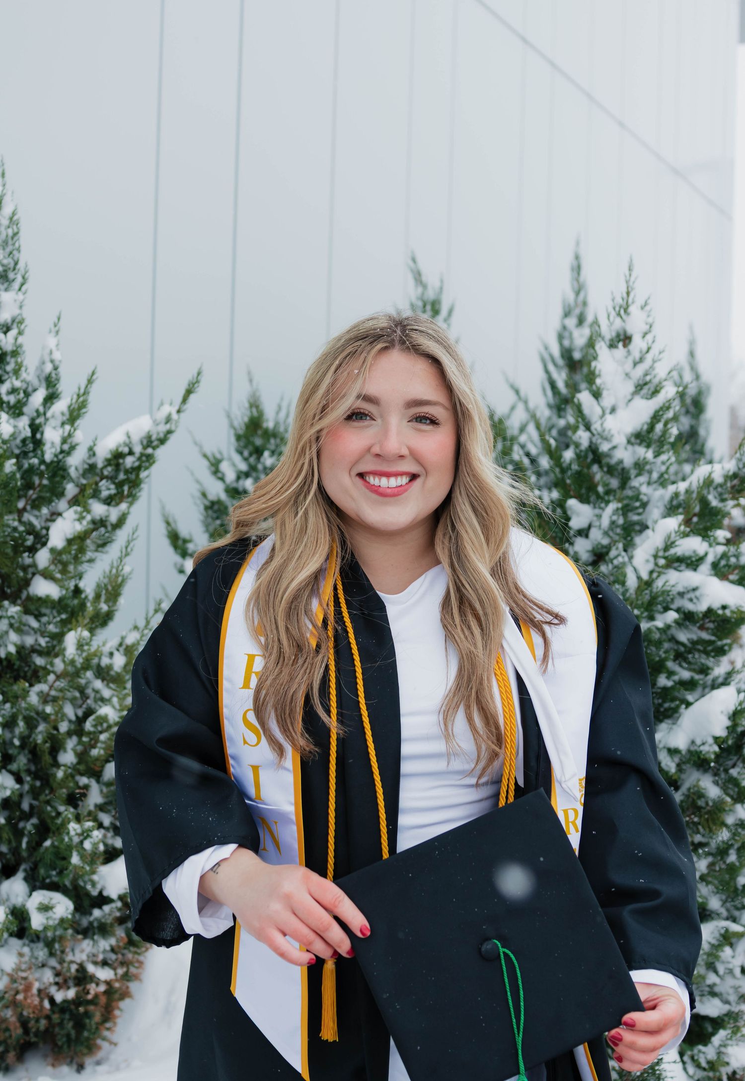 Smiling blonde graduate in cap and gown holding mortarboard near snow-dusted evergreen trees.