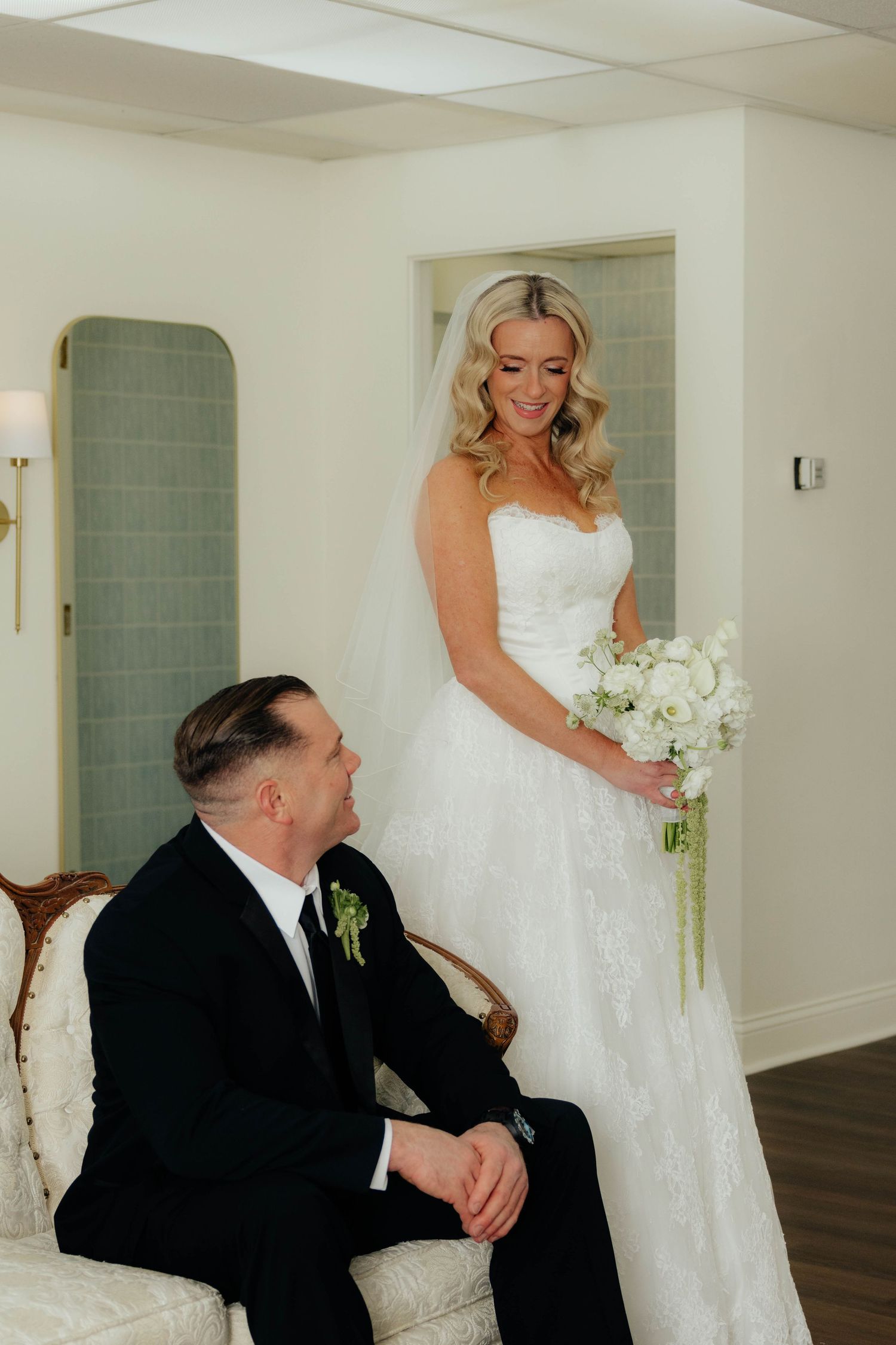 Bride in lace wedding gown smiling at groom seated in ornate chair during first look moment.