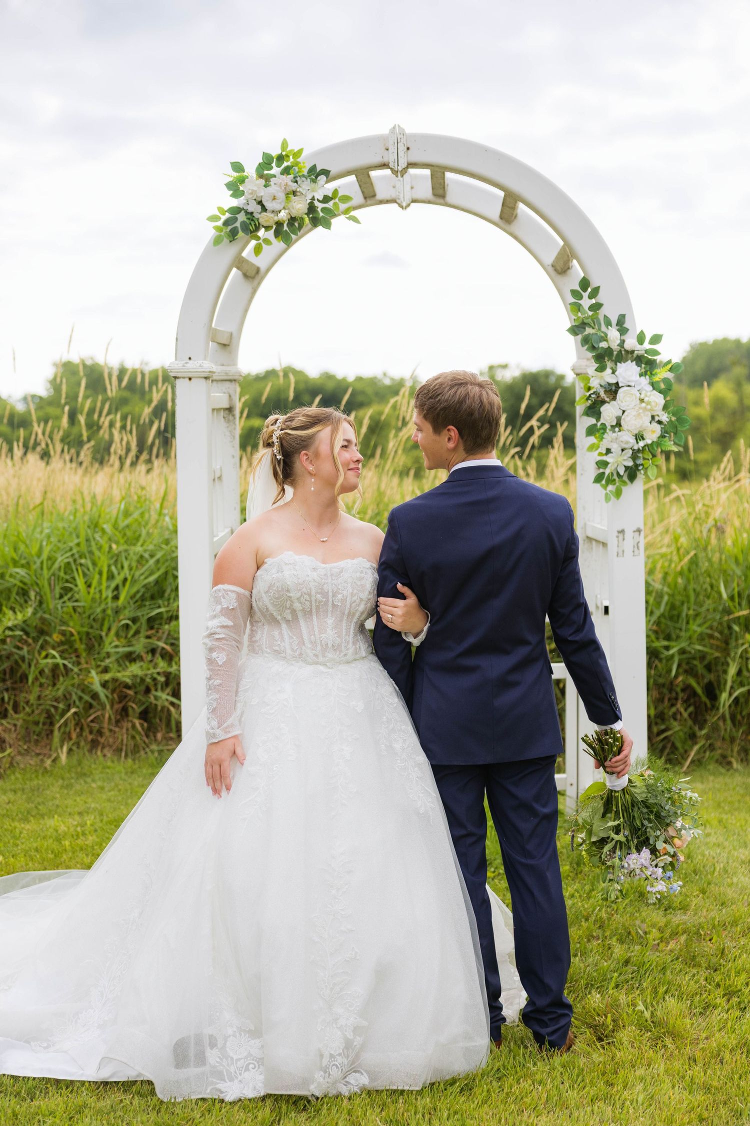 Bride in white gown and groom in navy suit pose under floral arch outdoors.