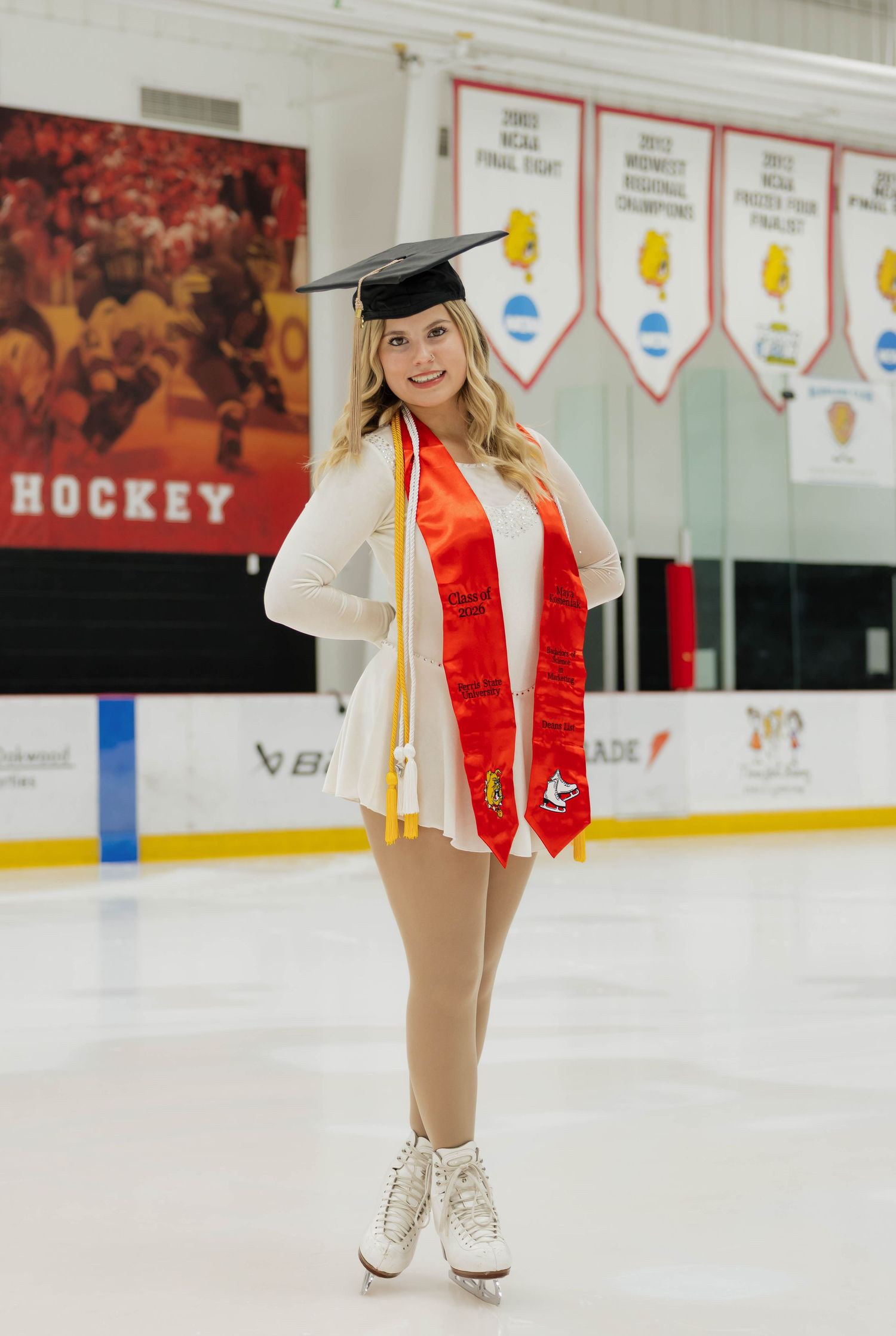 Graduate in cap and gown on ice rink wearing white skating dress and Ferris State red stole.