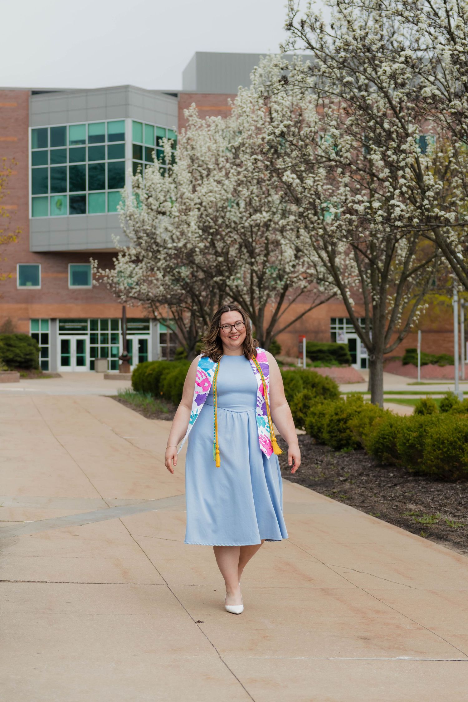 Graduate in light blue dress and colorful stole walking on campus pathway with blooming white trees behind.
