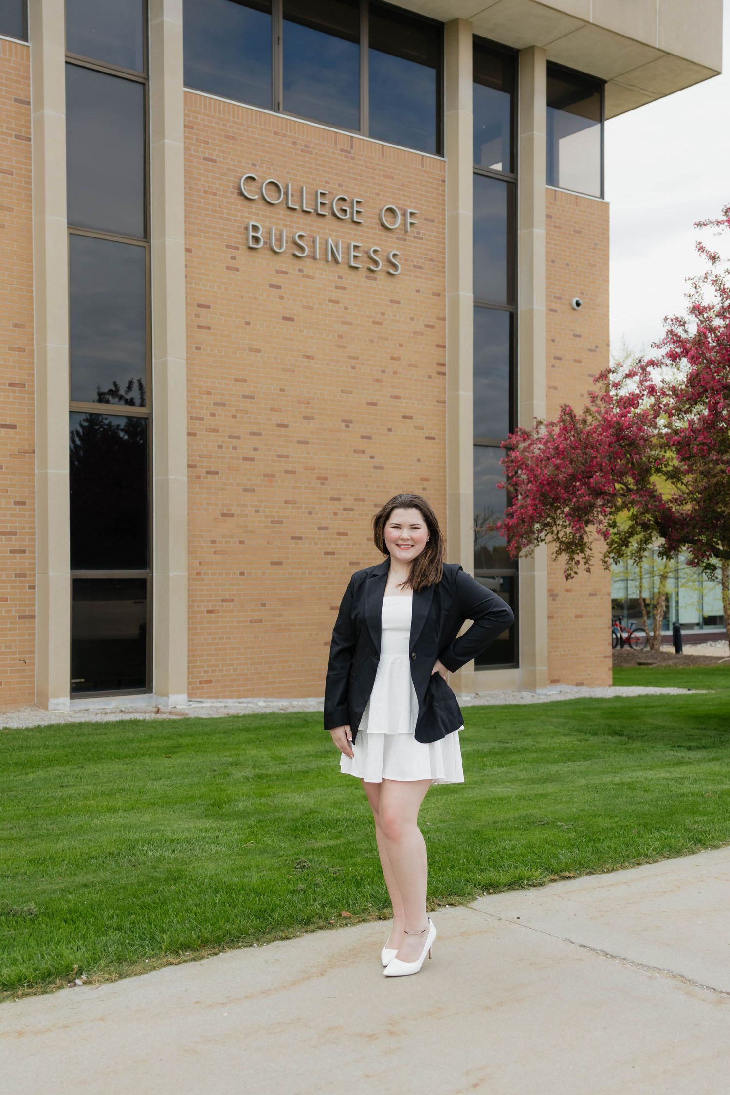 Female graduate in white dress and black blazer posing in front of College of Business building.