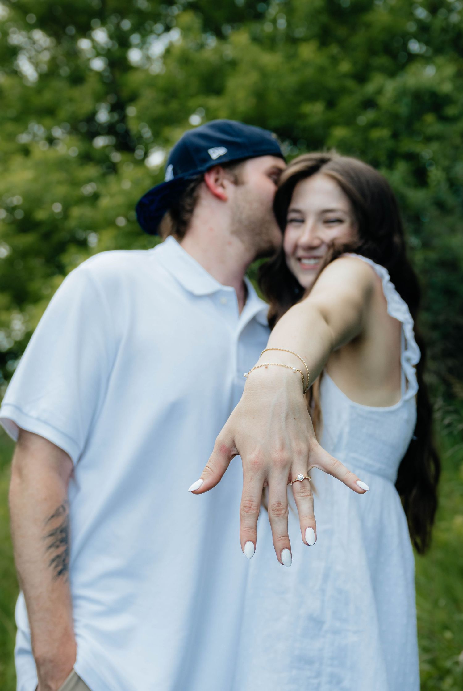 Smiling couple outdoors, woman extending hand to display engagement ring while partner kisses her cheek.
