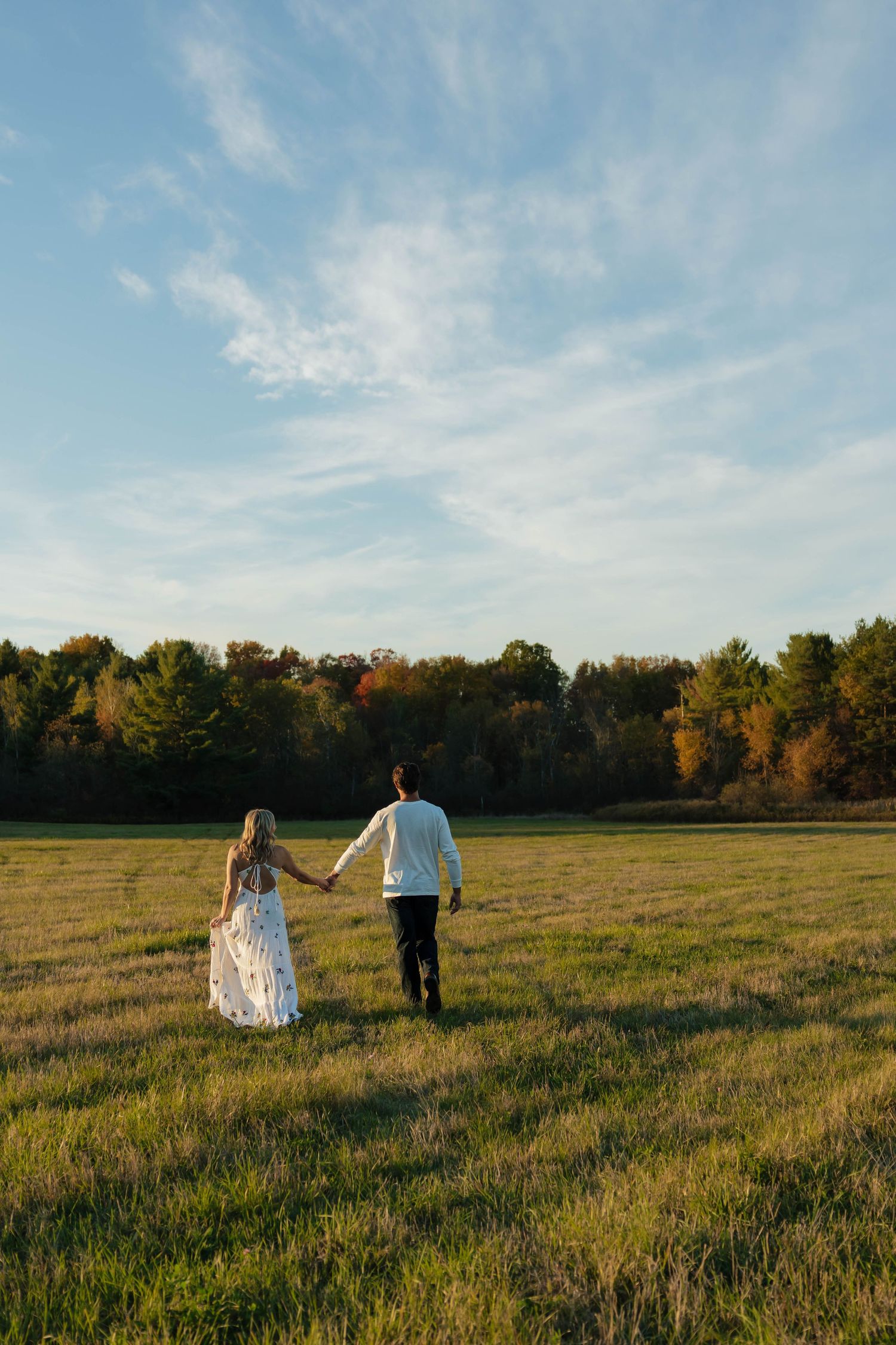 Couple holding hands walking through a vast green field toward autumn trees under a blue sky.
