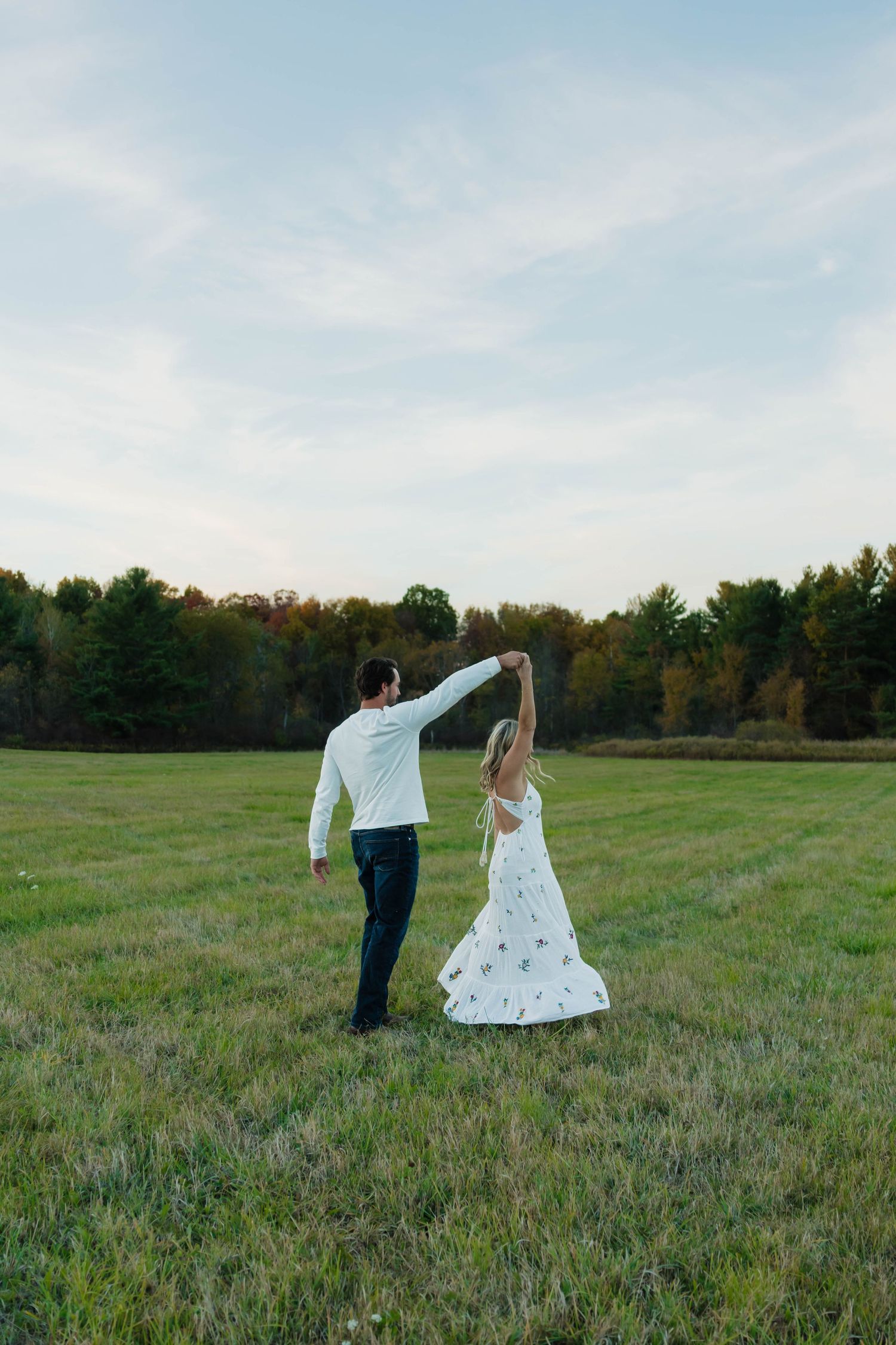 Couple dancing together in an open green field with autumn trees in the background at dusk.