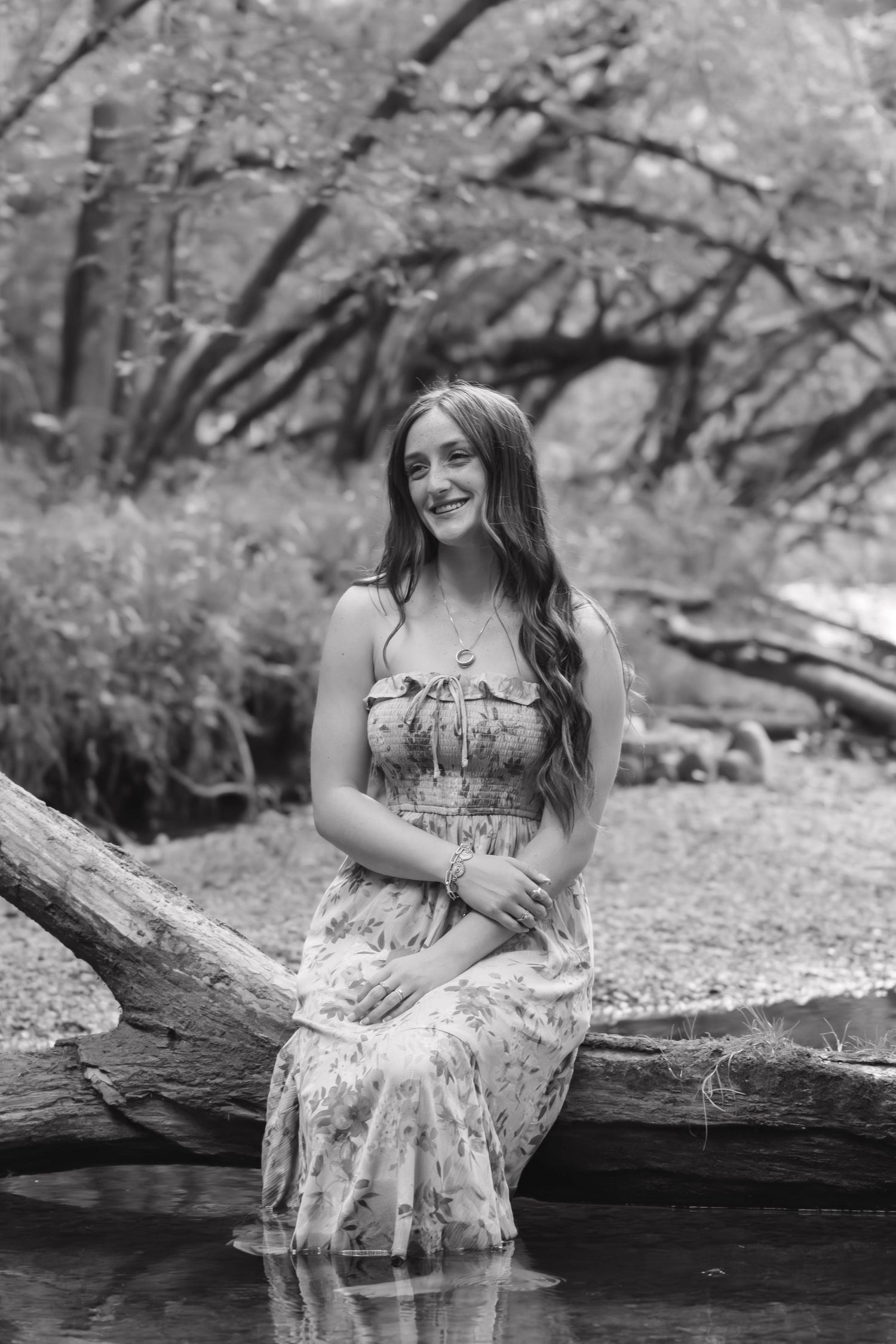 Black and white portrait of a teenage girl in a floral dress sitting on a log by water.