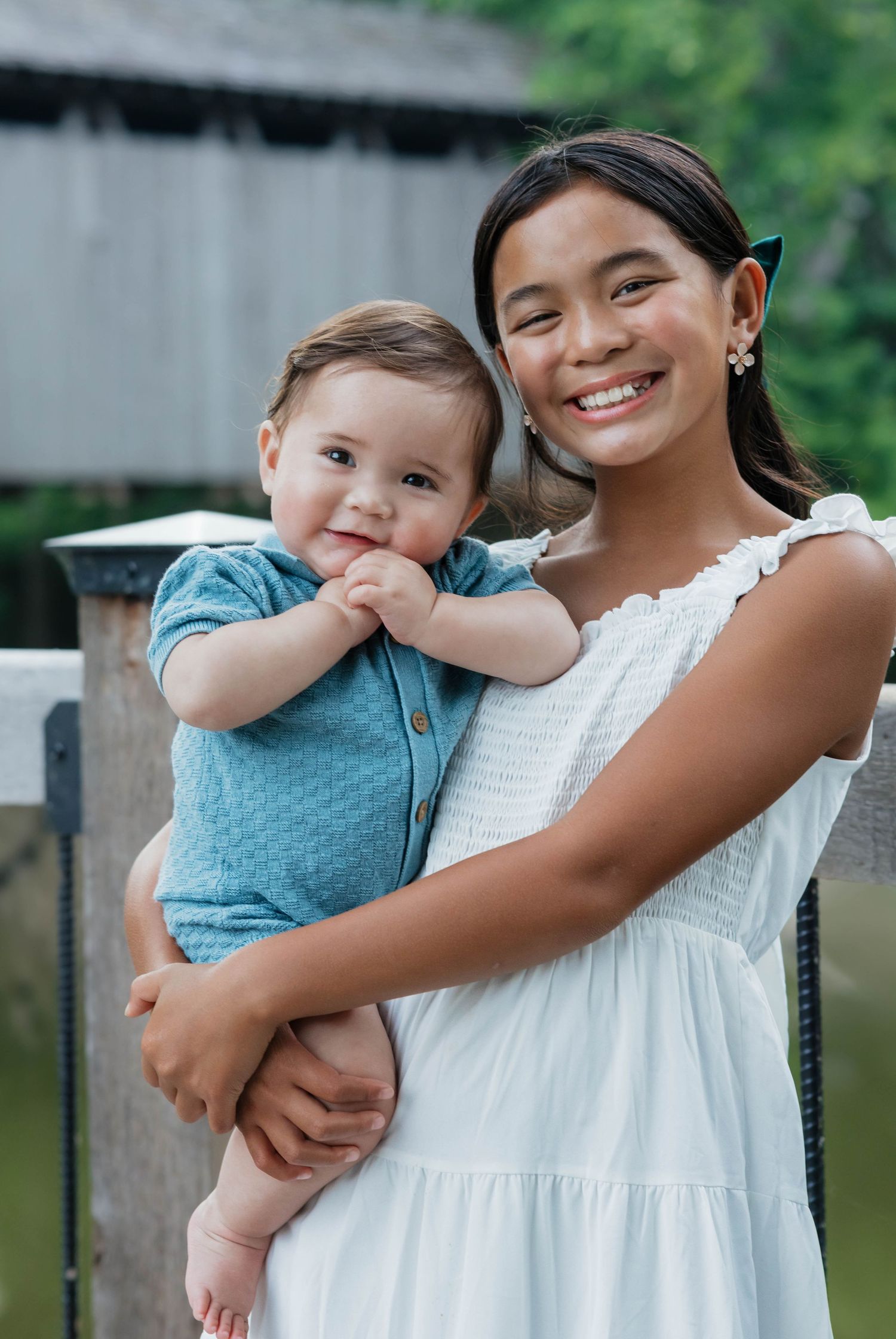 Smiling teenage girl holding an adorable baby boy outdoors near a bridge.