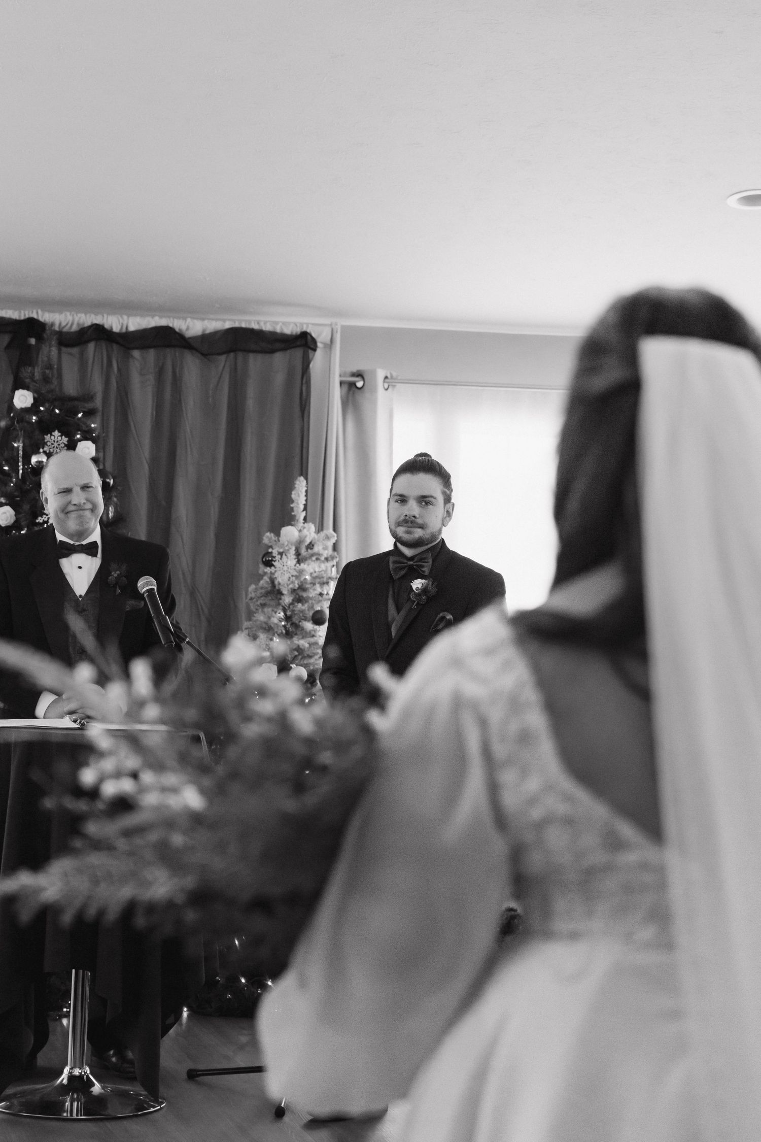 Black and white photo of groom watching bride walk down the aisle at indoor winter wedding ceremony.