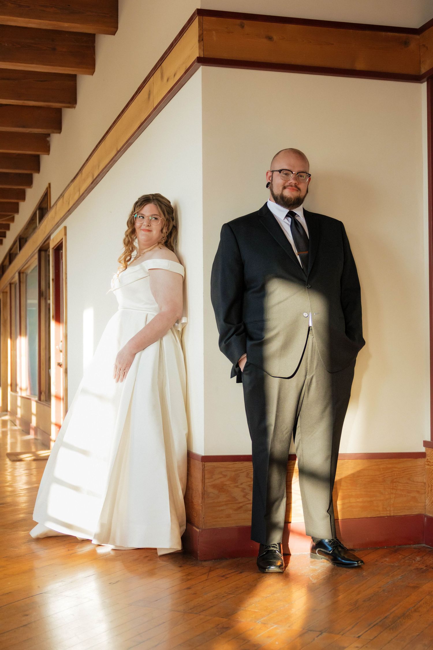 Bride in off-shoulder white gown and groom in dark suit pose in sunlit wooden hallway.