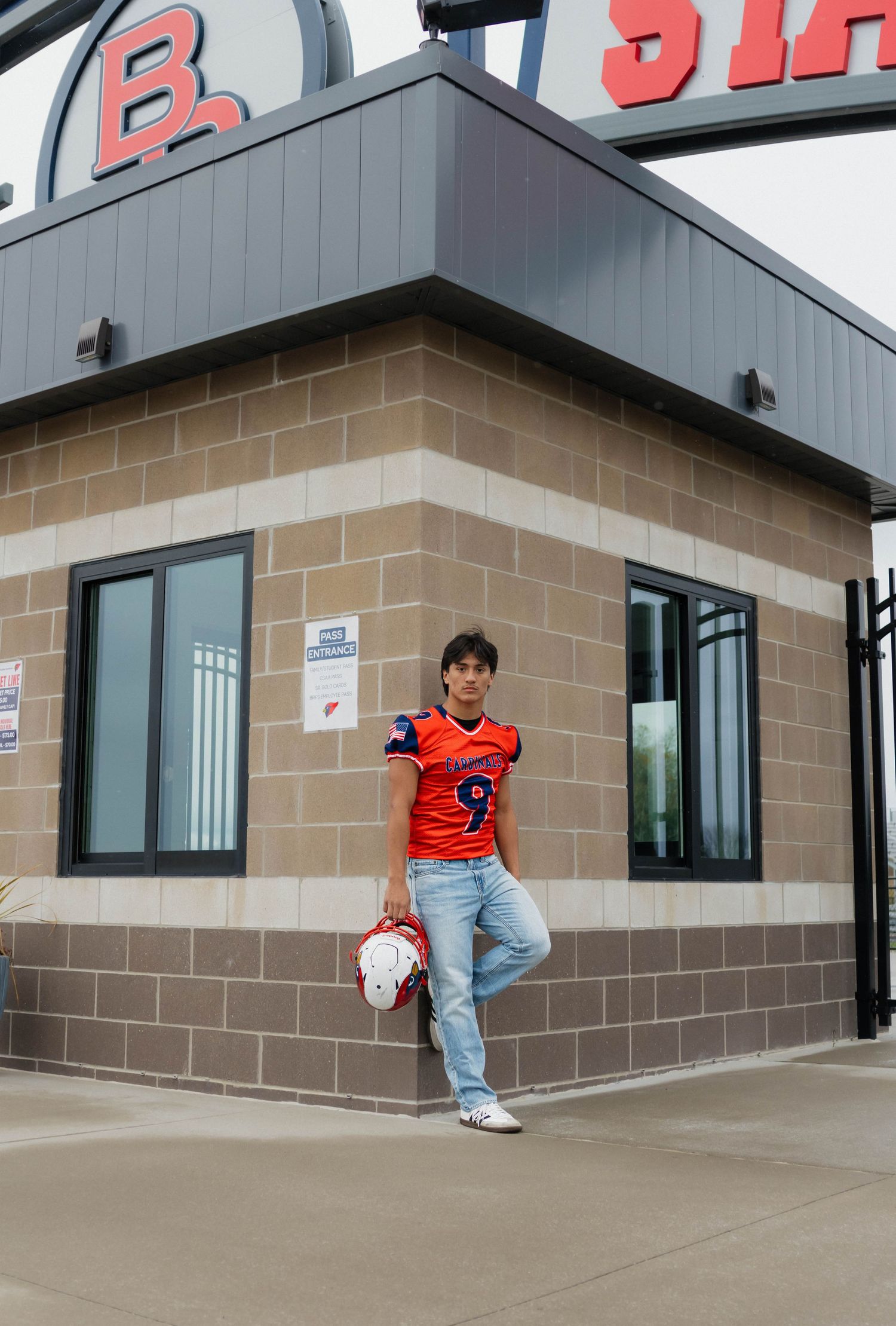 Teen football player in orange Cardinals jersey #9 holds helmet outside stadium entrance.
