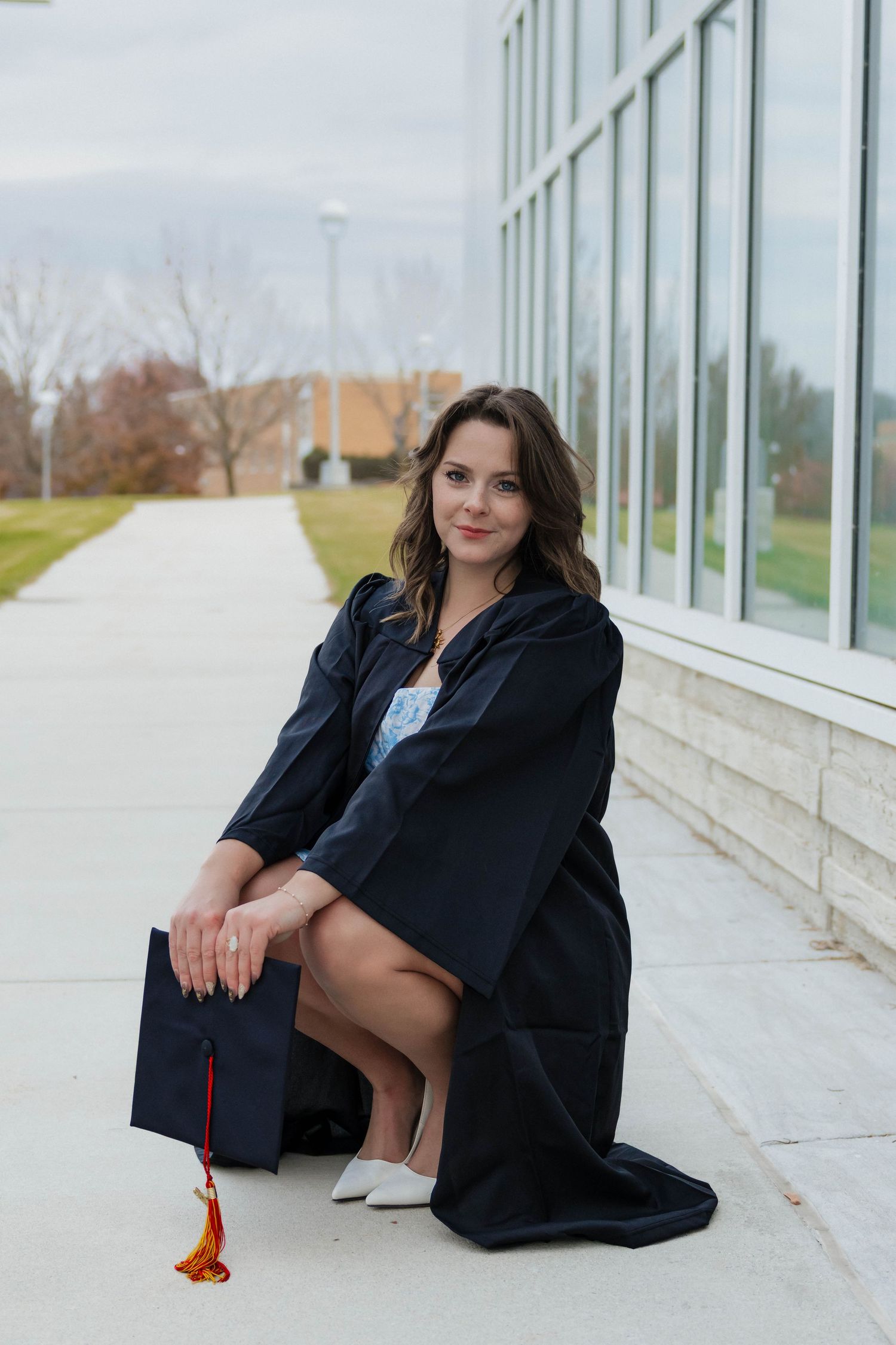 Female graduate in black gown sitting on campus walkway holding graduation cap with colorful tassel.