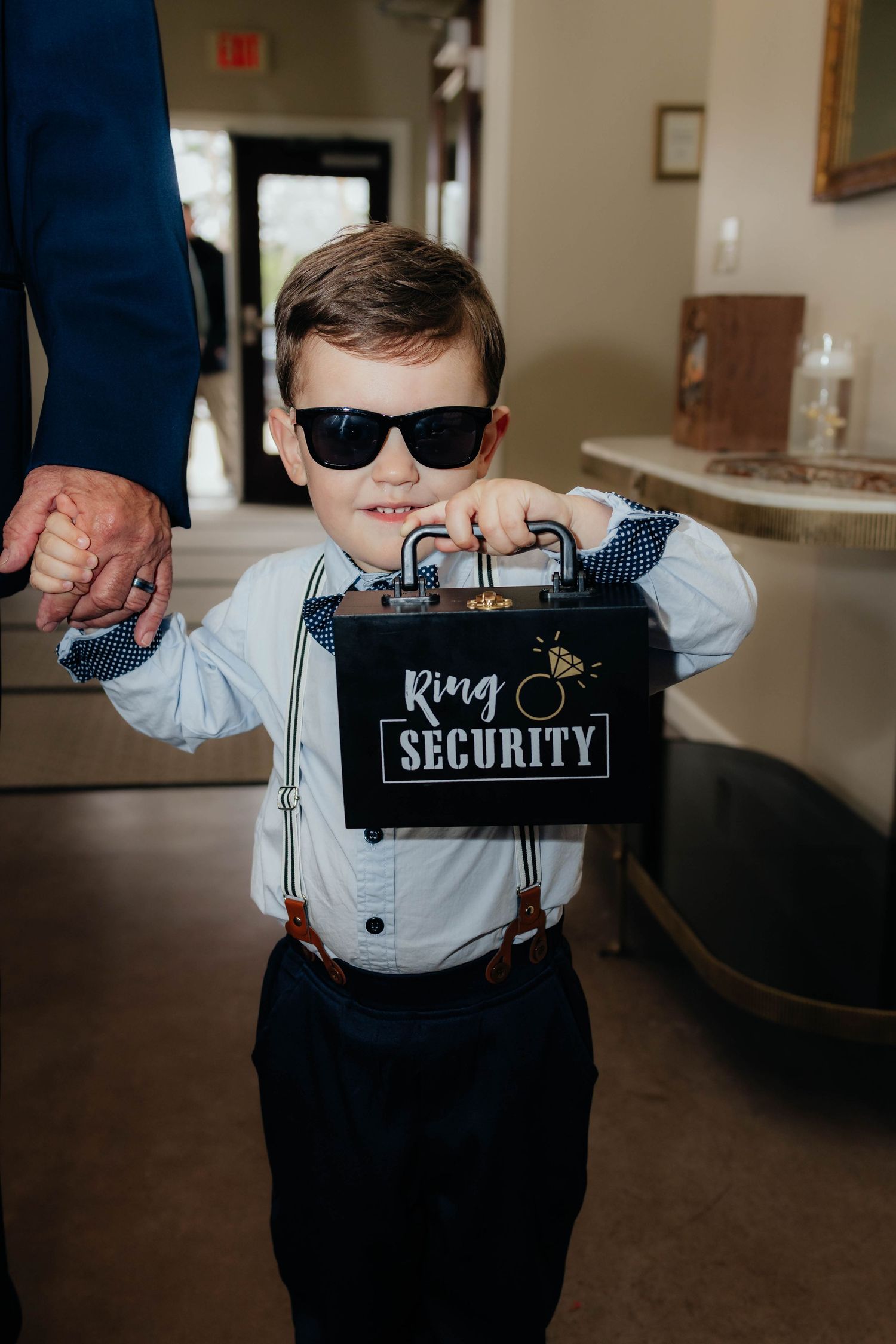 Adorable ring bearer in suspenders holds Ring Security briefcase and wears sunglasses at wedding ceremony.