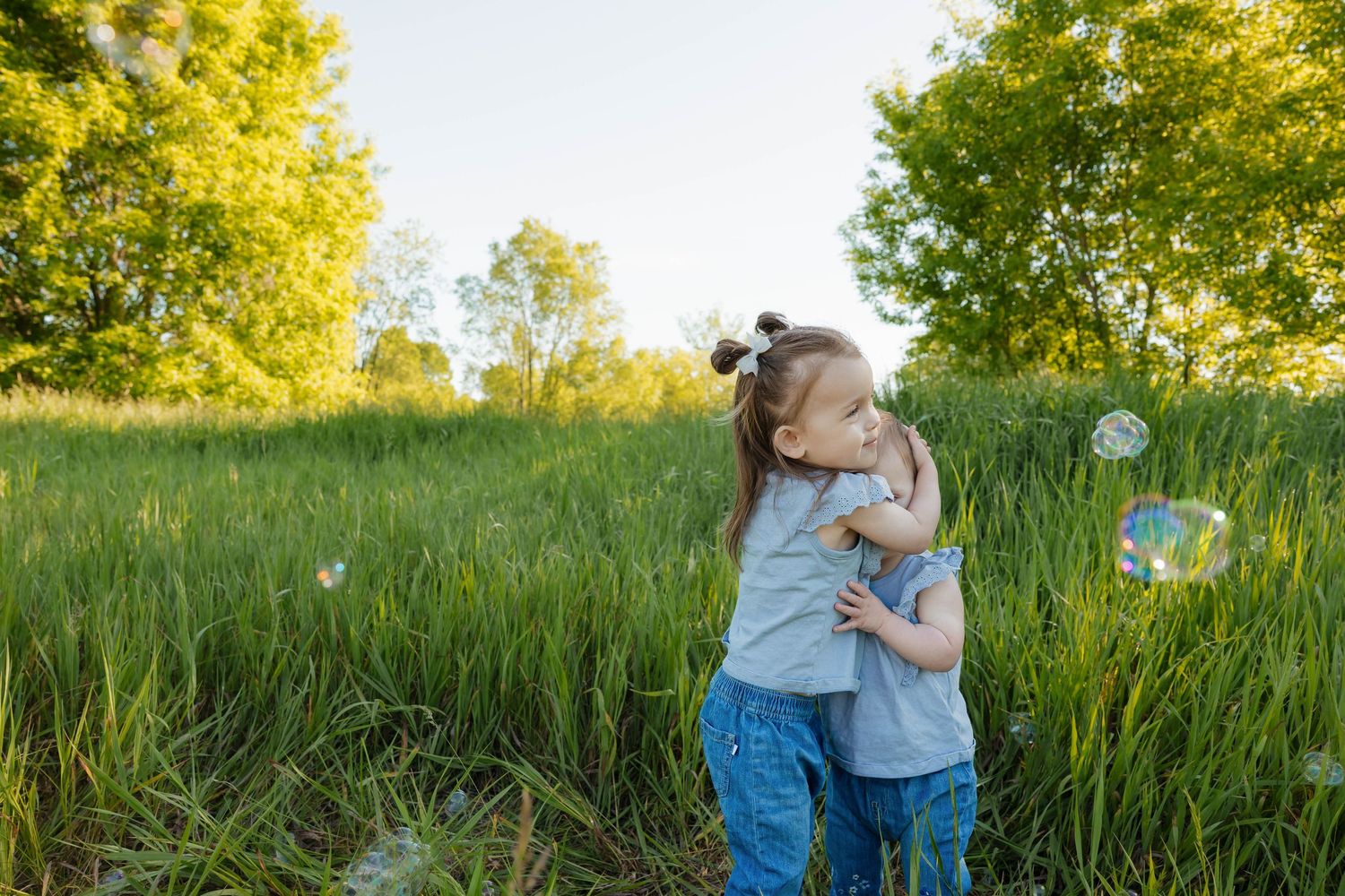 Two young girls hugging in a lush green field with soap bubbles floating around them on a sunny day.