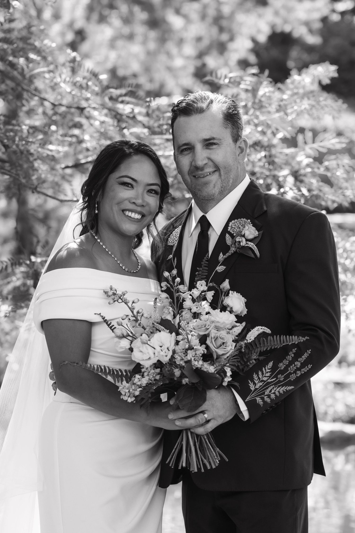 Black and white portrait of smiling bride and groom holding a floral bouquet outdoors.