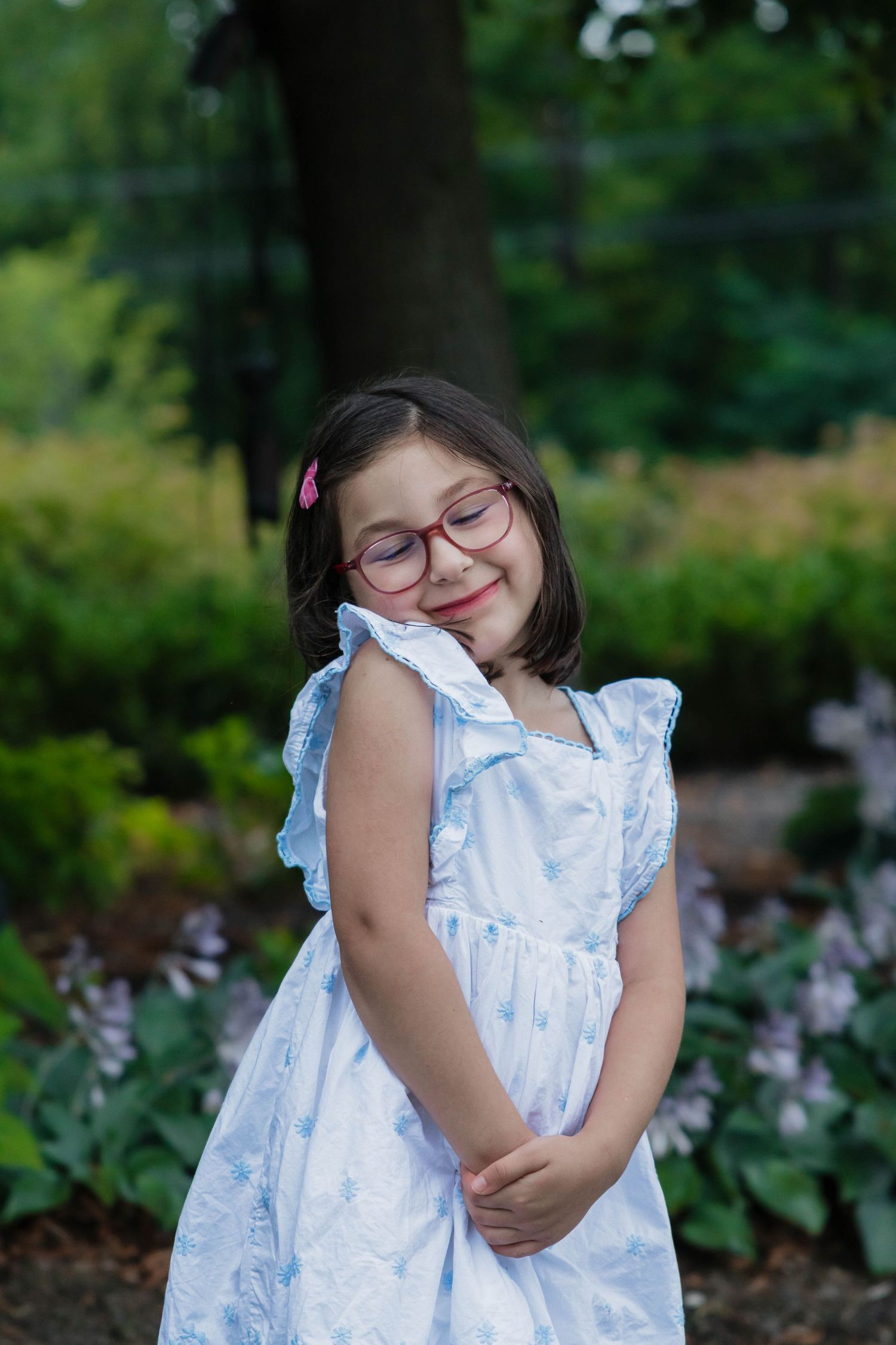 Young girl in blue floral dress with pink glasses smiles shyly in lush garden setting.