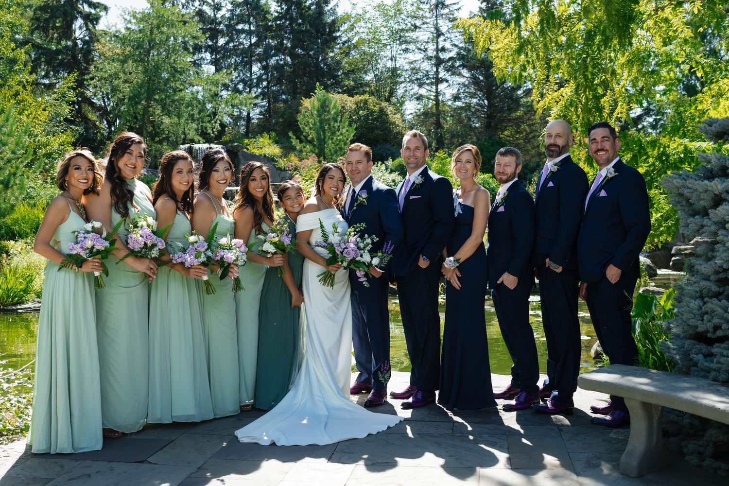 Wedding party poses together with bridesmaids in sage green gowns and groomsmen in navy suits in garden.