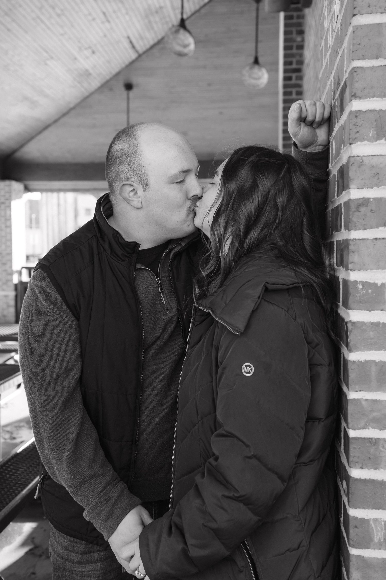 Black and white photo of a couple sharing a kiss against a brick wall in winter.