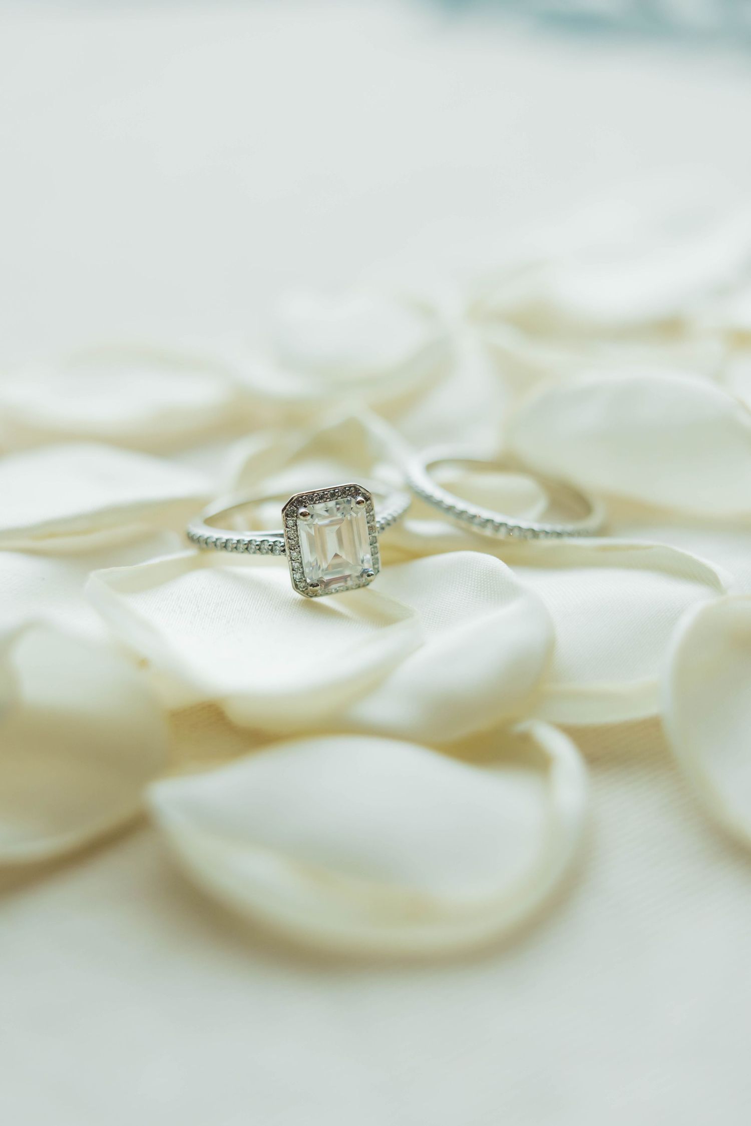 Emerald-cut diamond engagement ring with halo setting and wedding band resting on white rose petals.