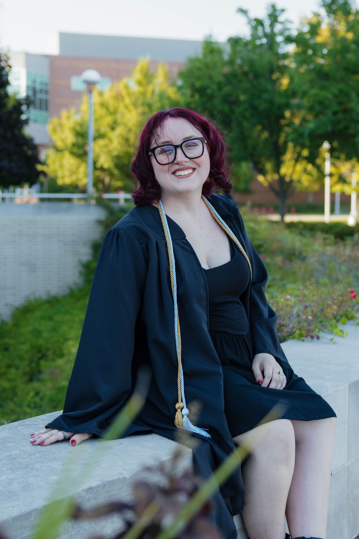 Happy female graduate in black gown with honor cords sitting outdoors on campus wall on a sunny day.