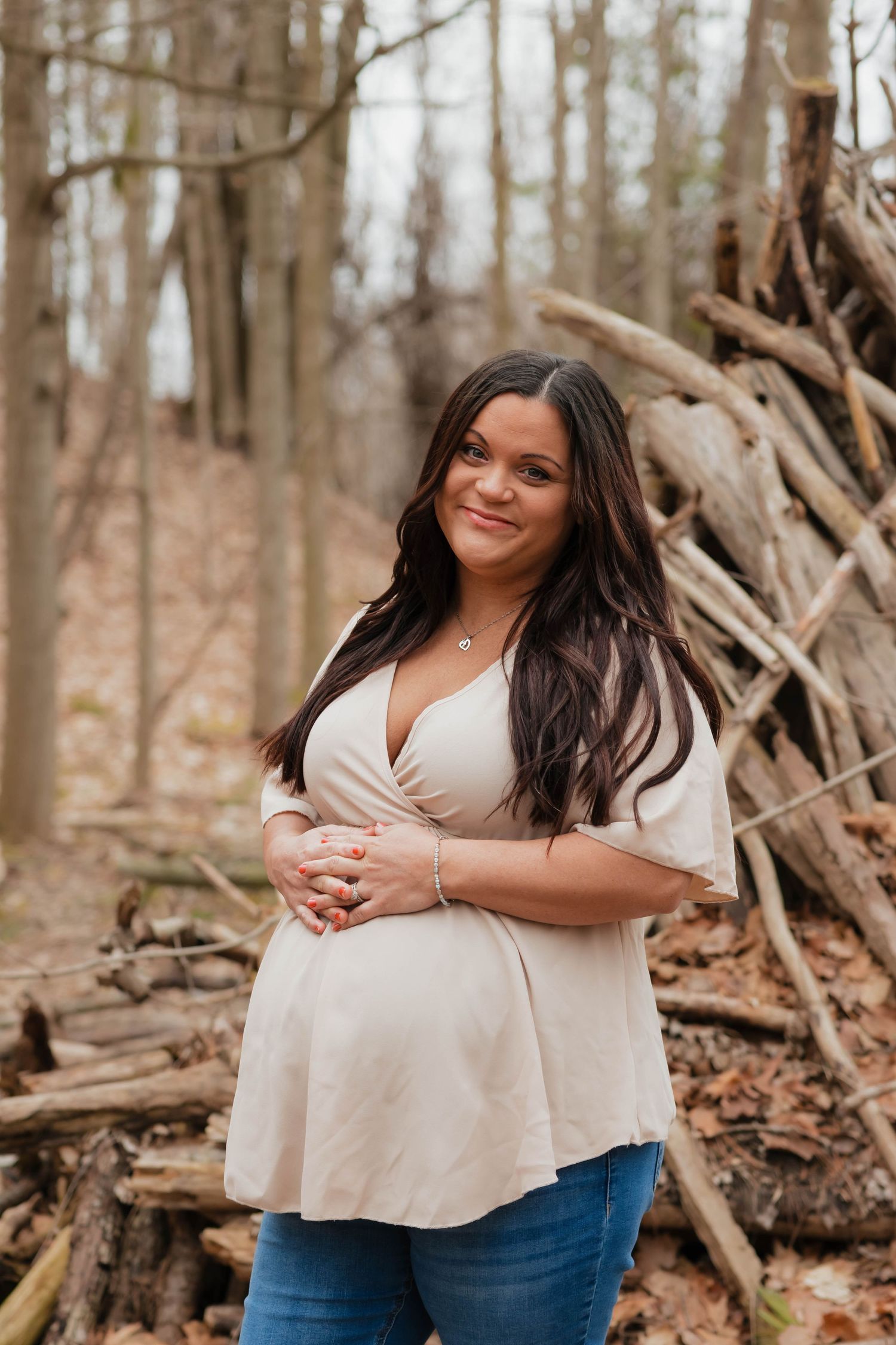 Pregnant woman in beige top and jeans smiling in a forest setting with stacked logs behind her.