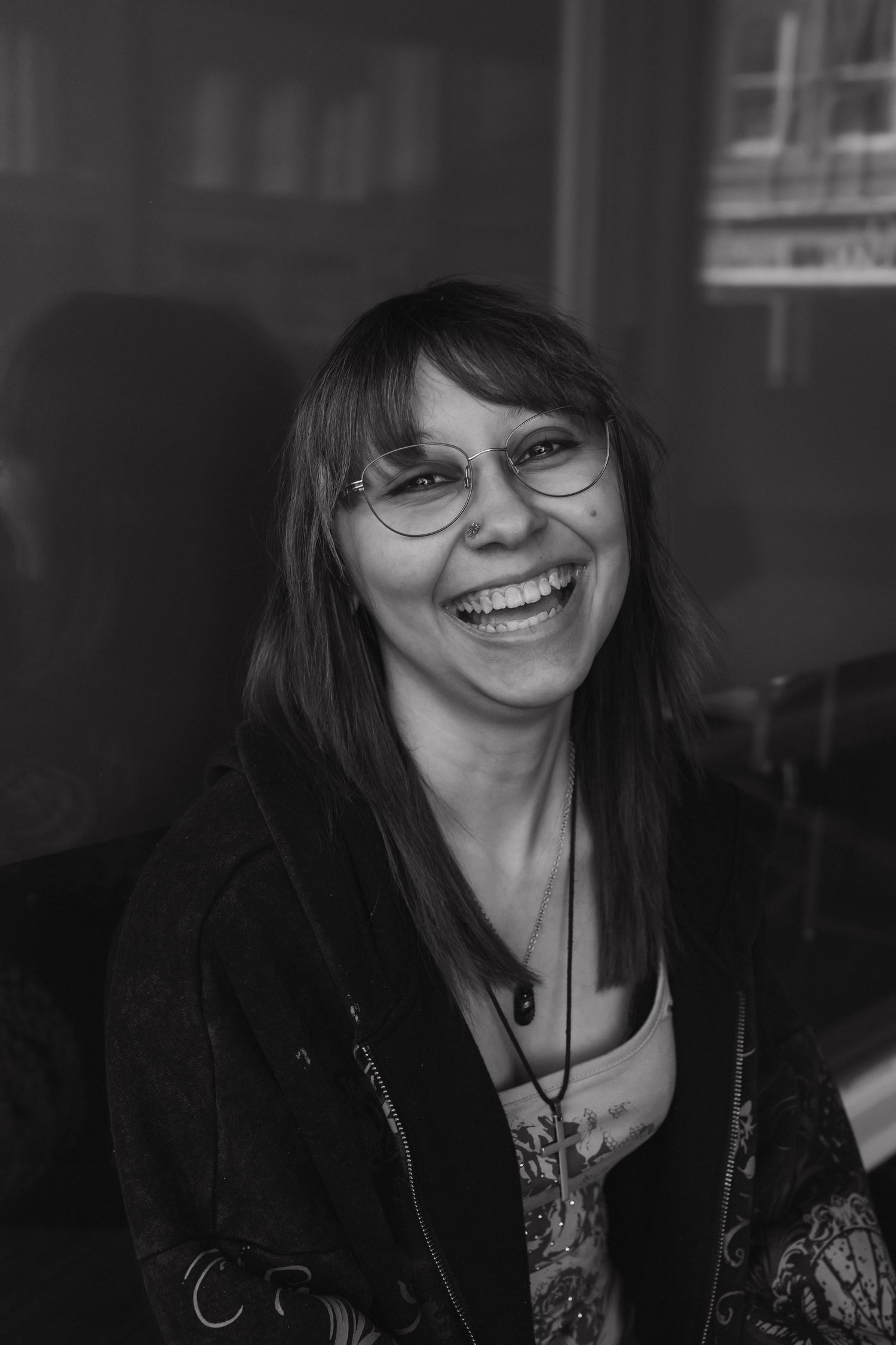 Black and white portrait of a laughing woman with glasses and long hair wearing a necklace and dark jacket.