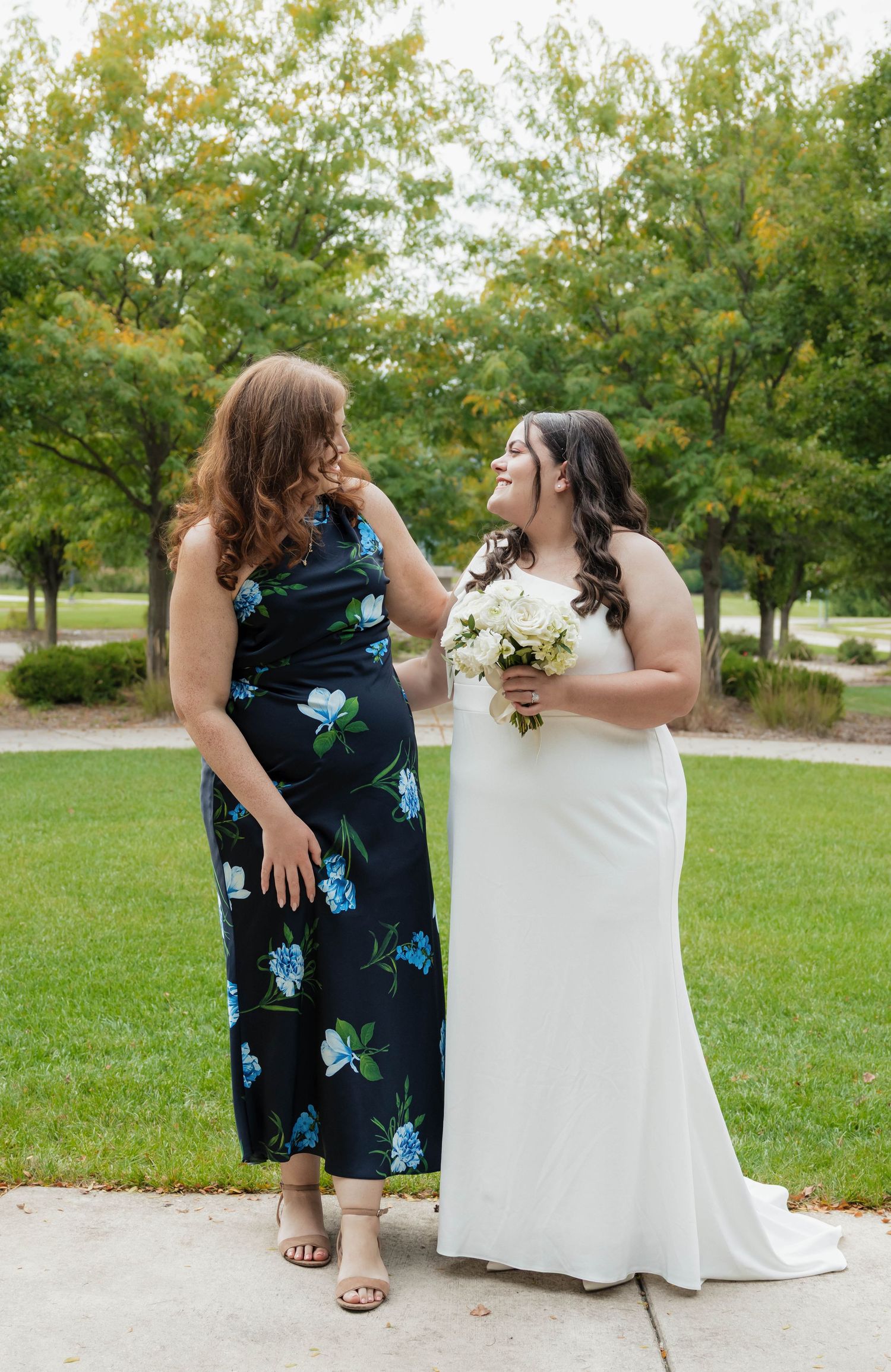Bride in white gown holding bouquet shares joyful moment with woman in floral dress outdoors.