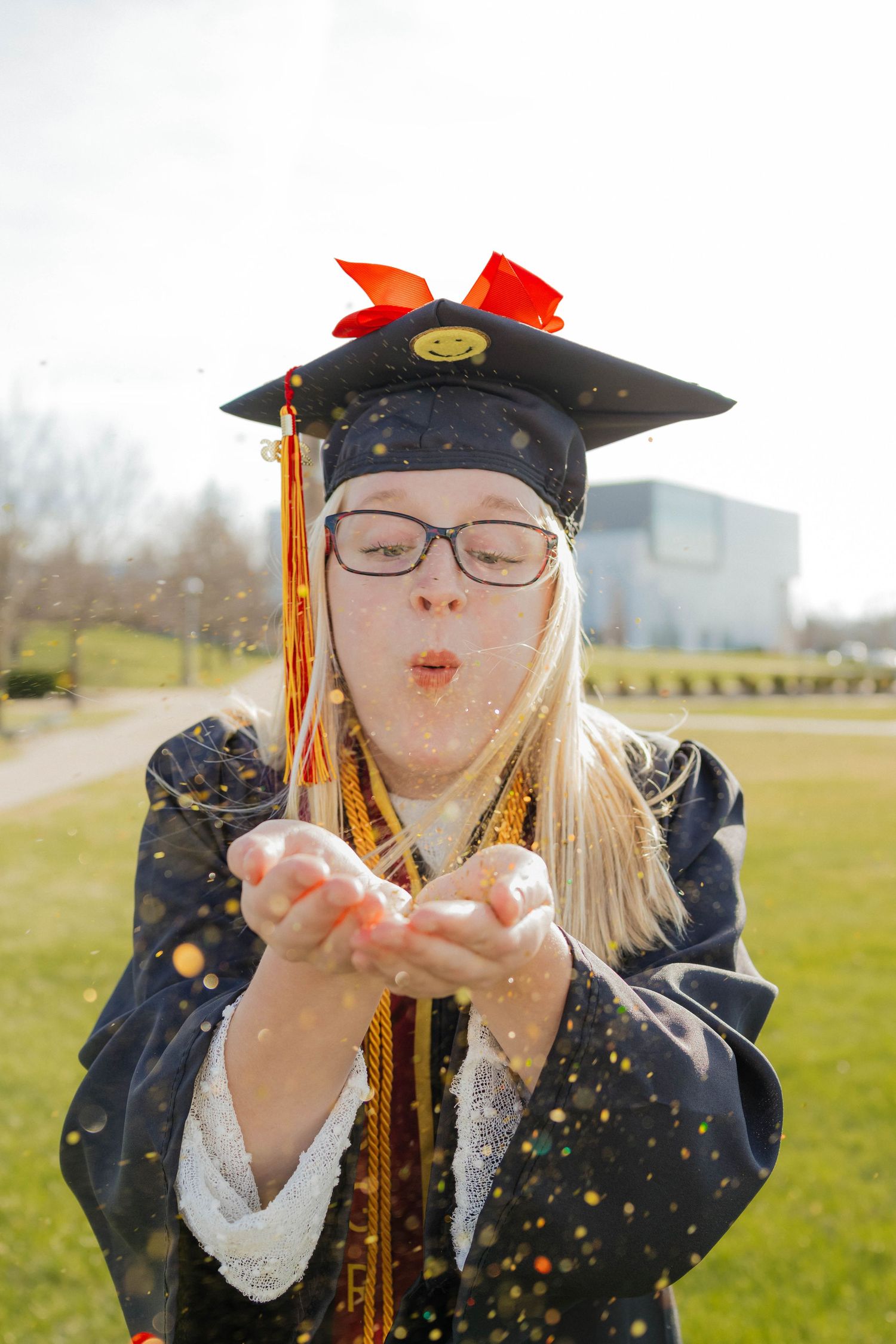 Blonde graduate in cap and gown blowing colorful confetti from her hands outdoors on campus.