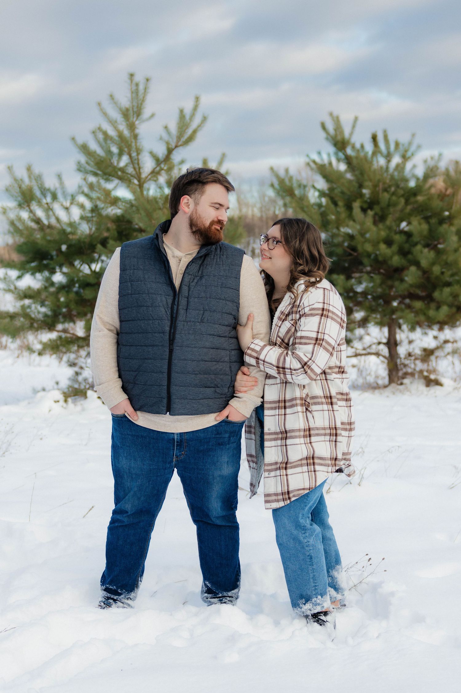 Couple smiling and gazing at each other while standing in an open snowy field with evergreen trees behind.