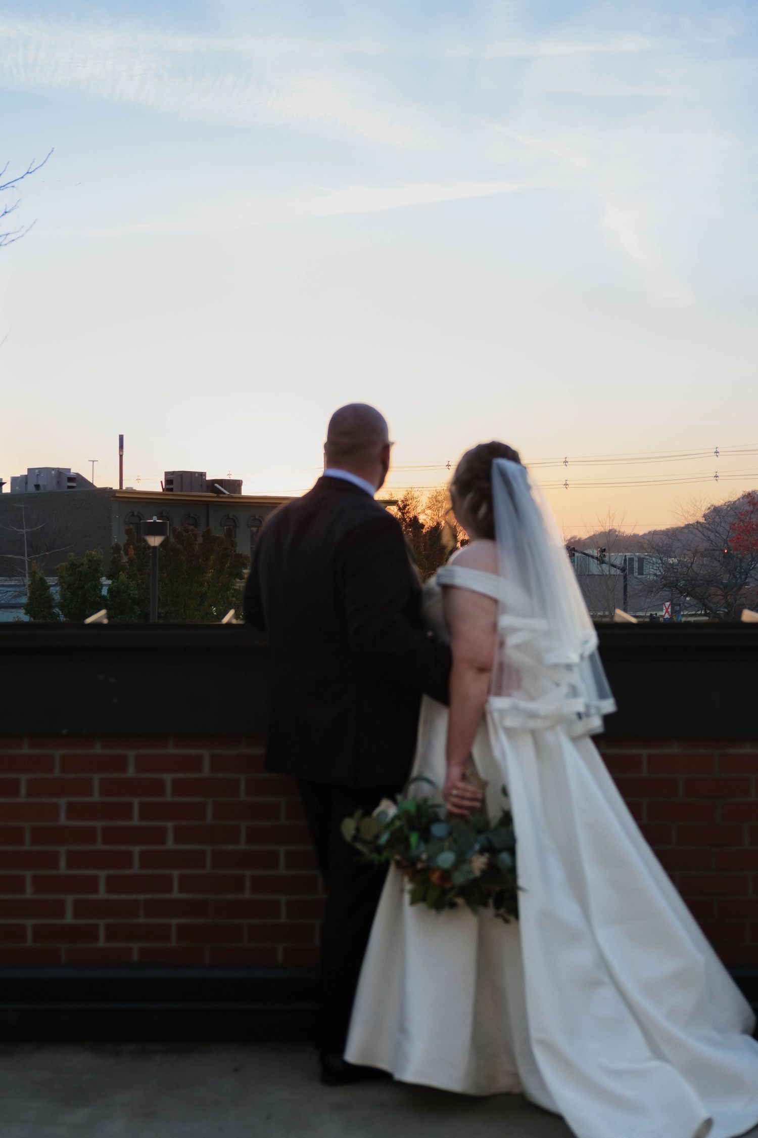 Bride and groom standing on rooftop terrace at sunset, bride in white gown with veil holding greenery bouquet.