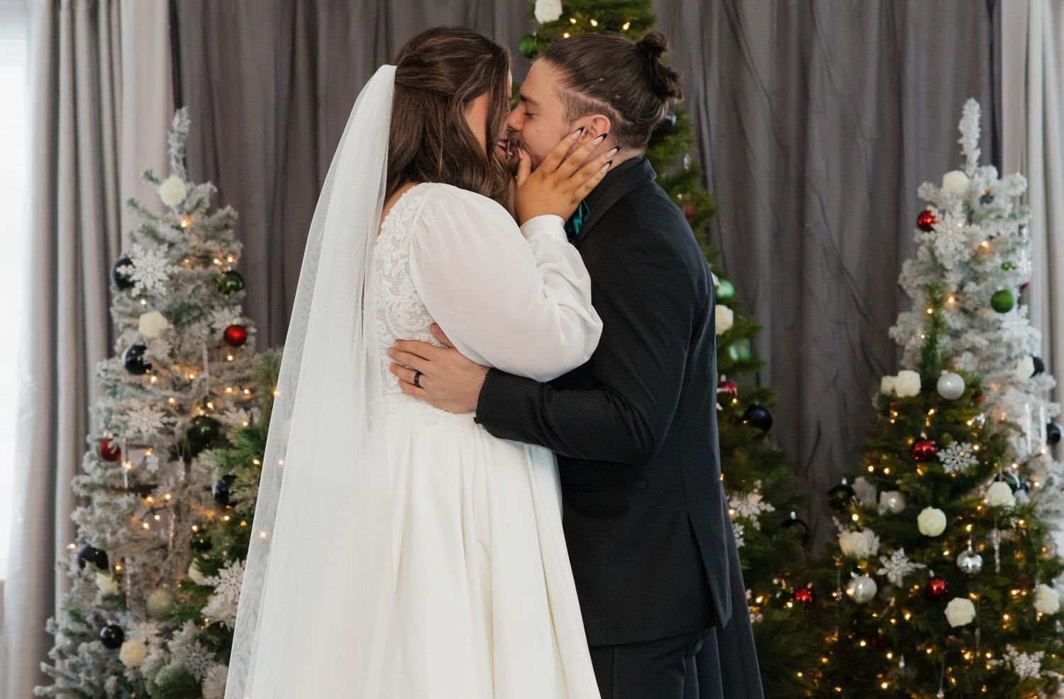 Bride and groom sharing a kiss surrounded by decorated Christmas trees indoors.