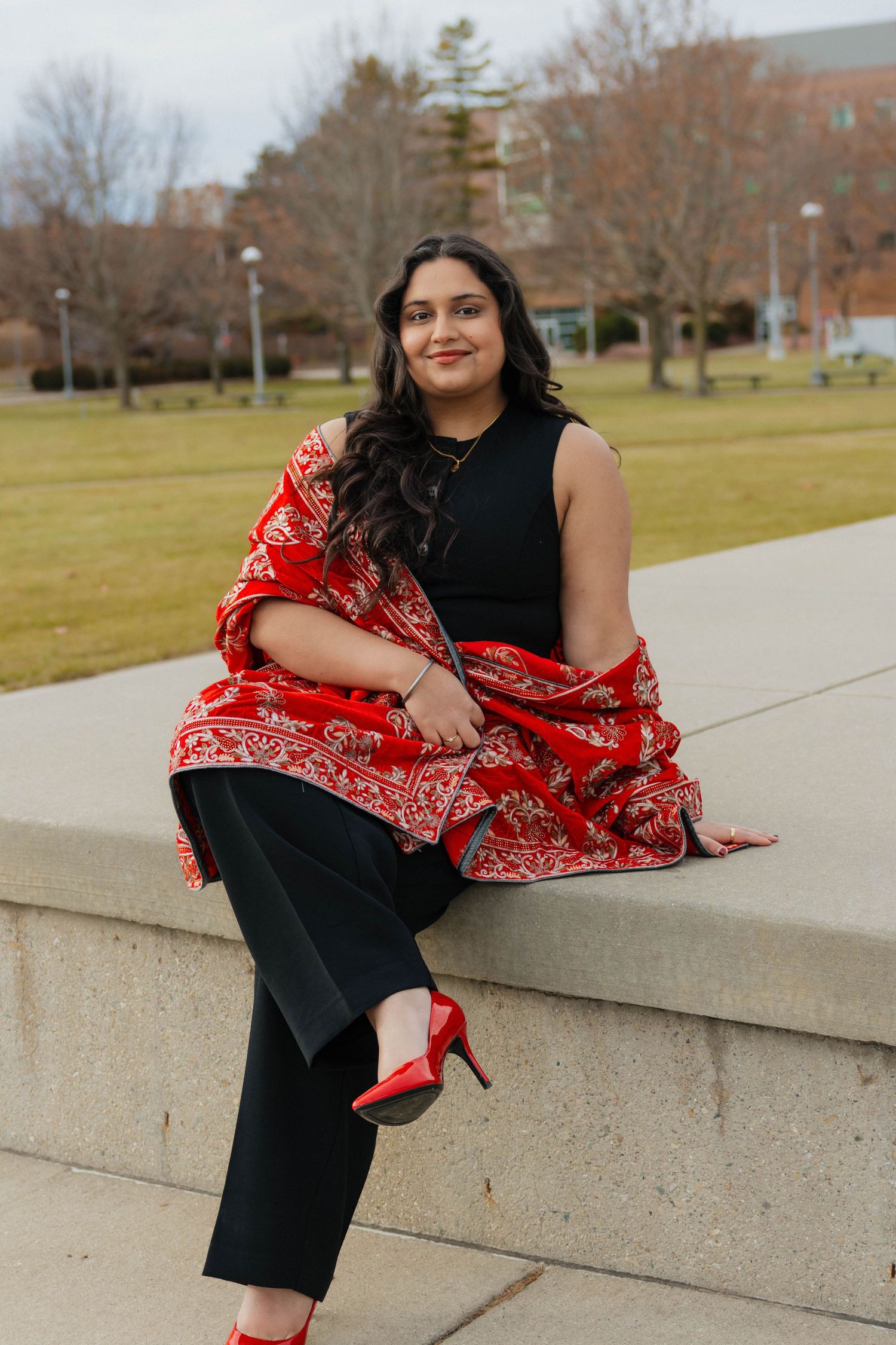 Young woman in black outfit with red paisley shawl and heels posing on a campus wall.