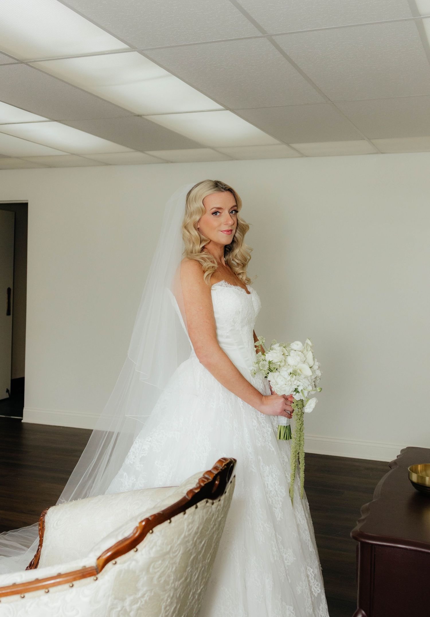 Elegant bride in strapless lace wedding gown with veil holding white bouquet in bridal suite.