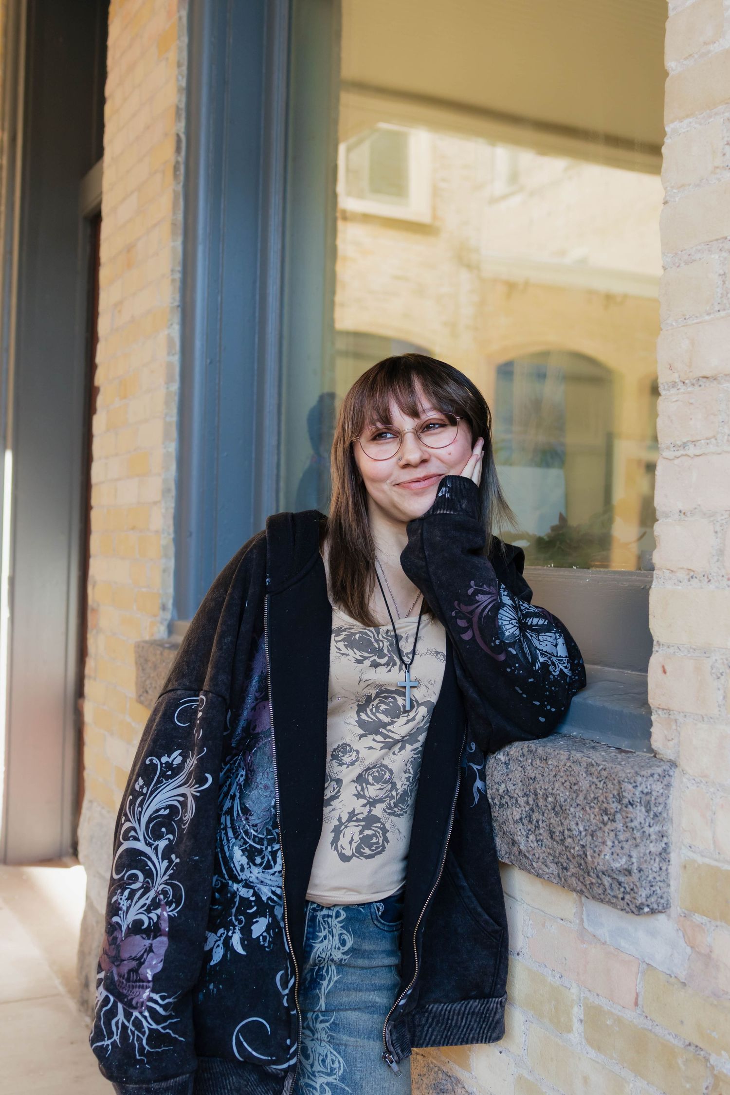 Woman with glasses leaning against a brick pillar outdoors, wearing a floral jacket and graphic tee.