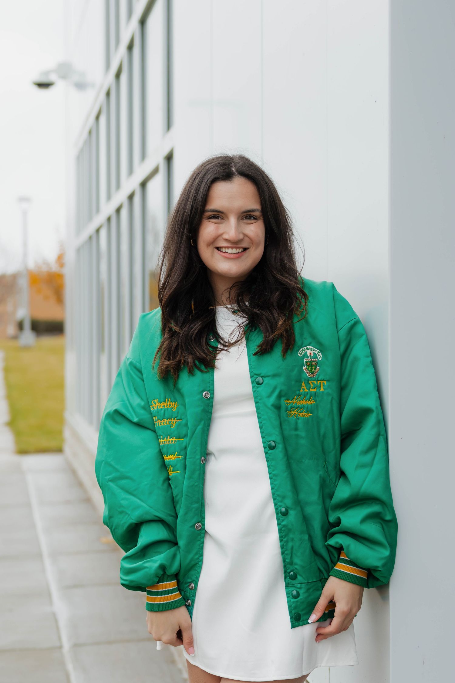 Young woman smiling in a green varsity jacket and white dress leaning against a building wall.