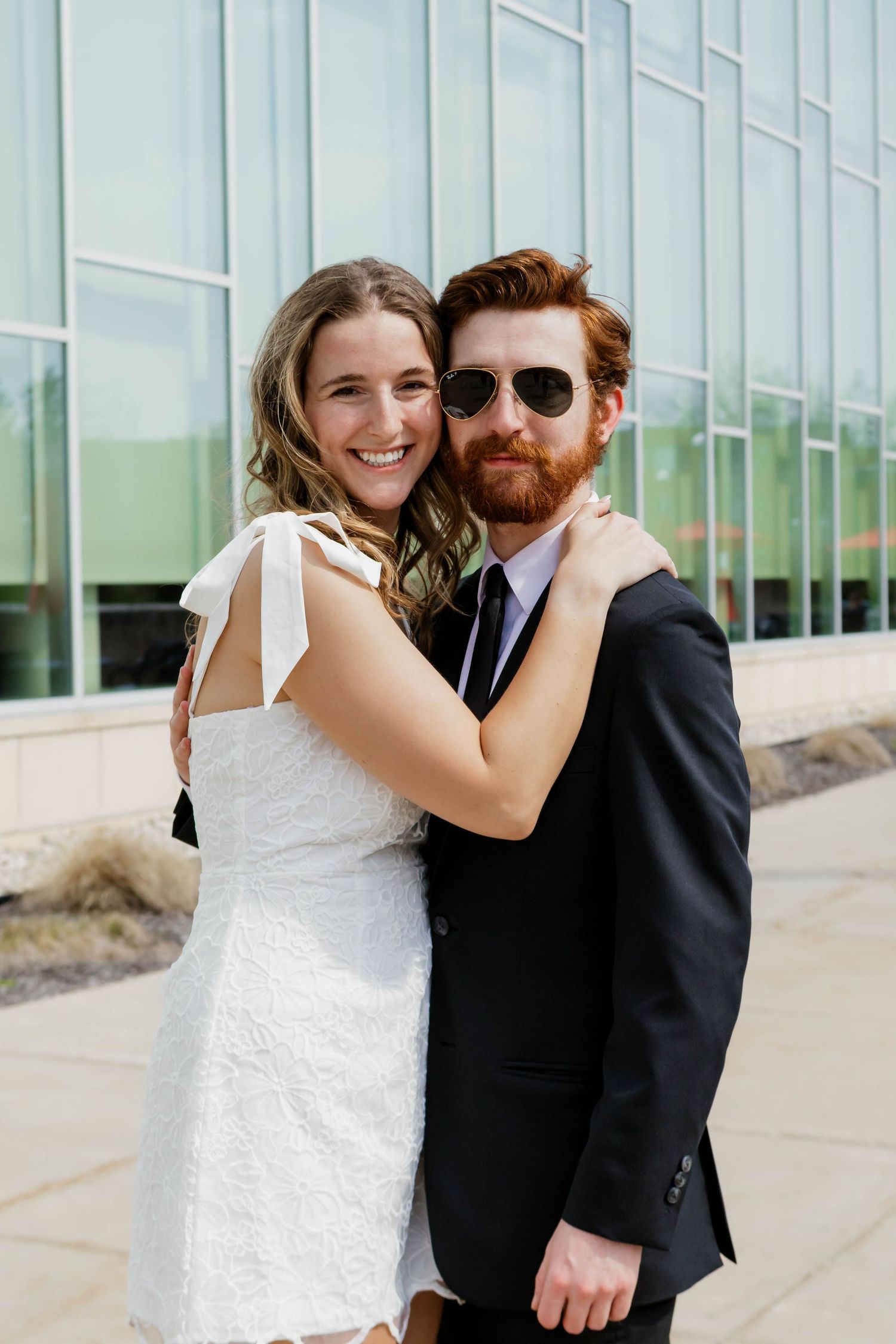 Happy couple embracing outdoors, woman in white dress and man in black suit with sunglasses.