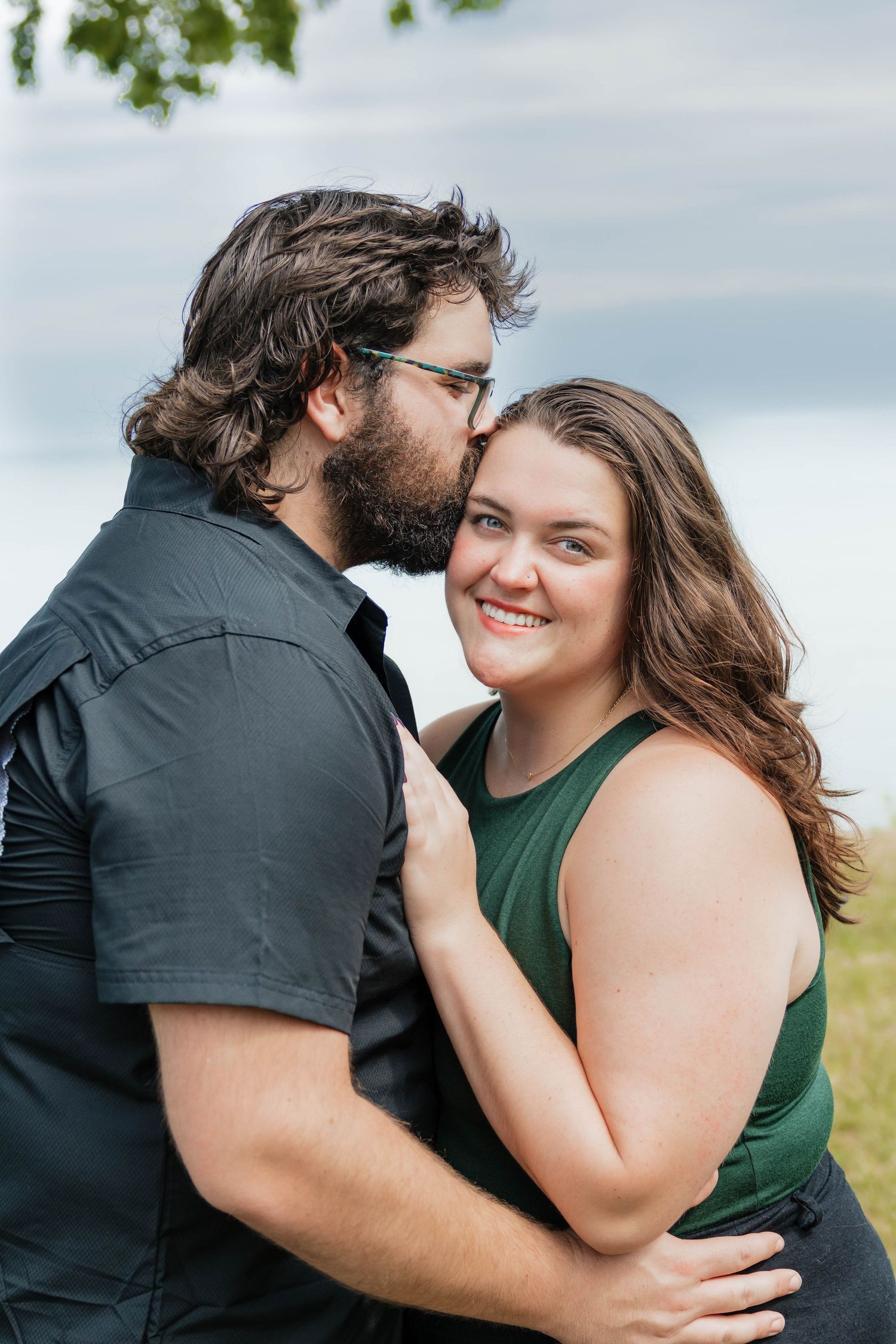 Couple sharing a tender kiss on the cheek during outdoor engagement photos with overcast sky backdrop.