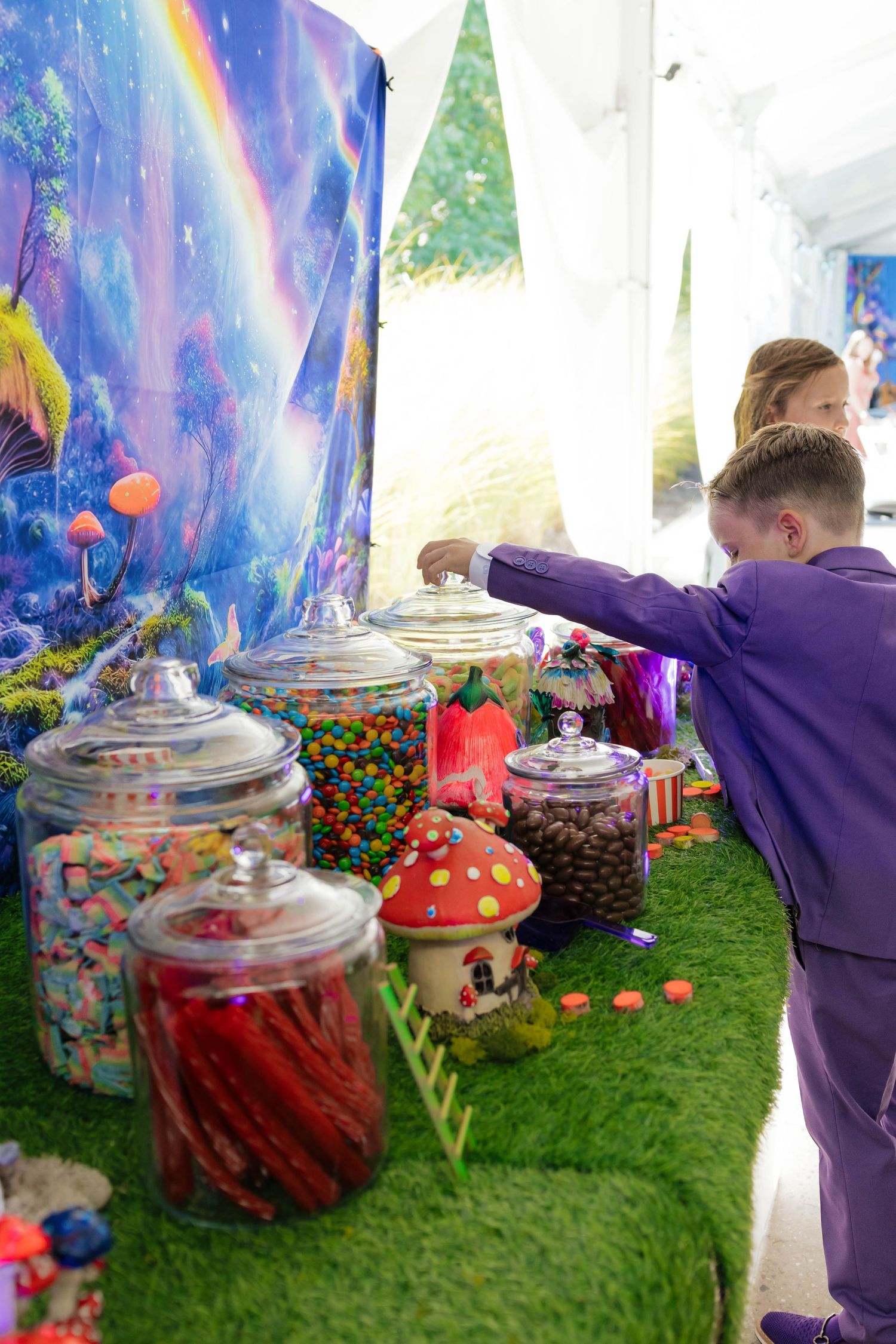 Child in purple suit reaches into candy jar at colorful fantasy-themed wedding candy buffet display.