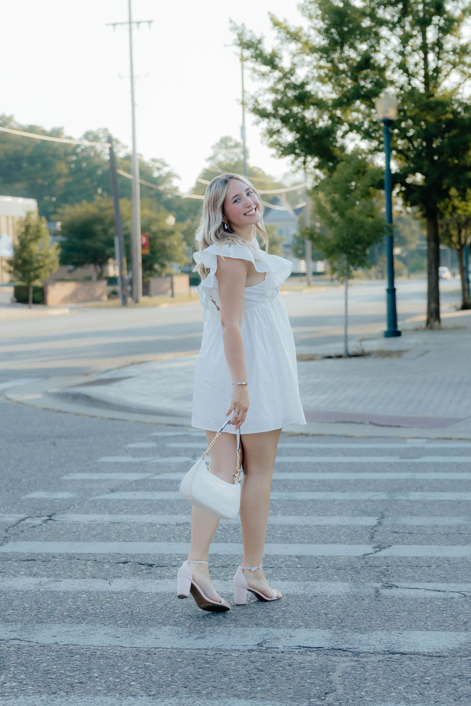Smiling blonde woman in white mini dress crossing street holding white purse wearing heels.