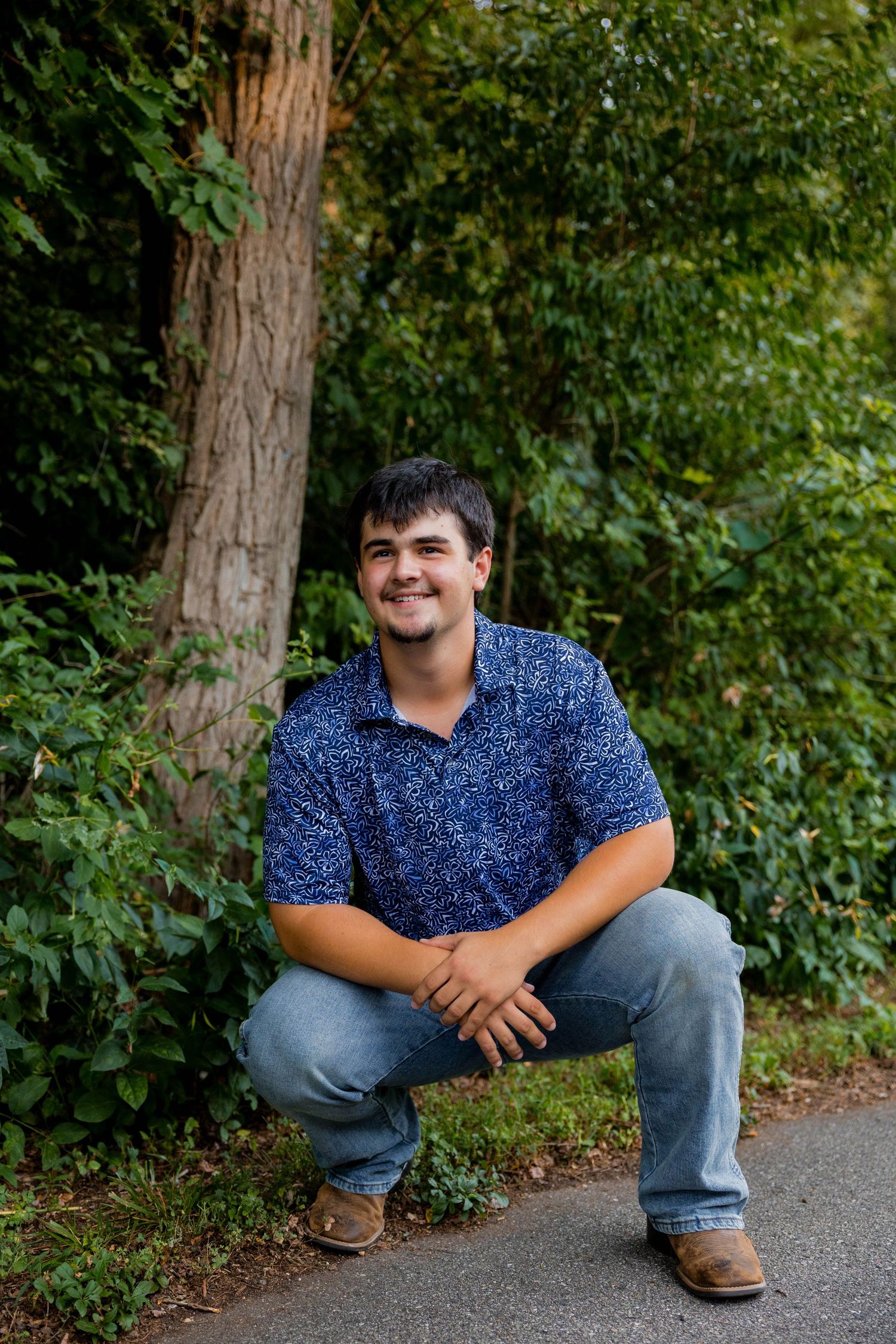 Young man in blue patterned shirt and jeans crouching outdoors near trees, smiling for a portrait photo.