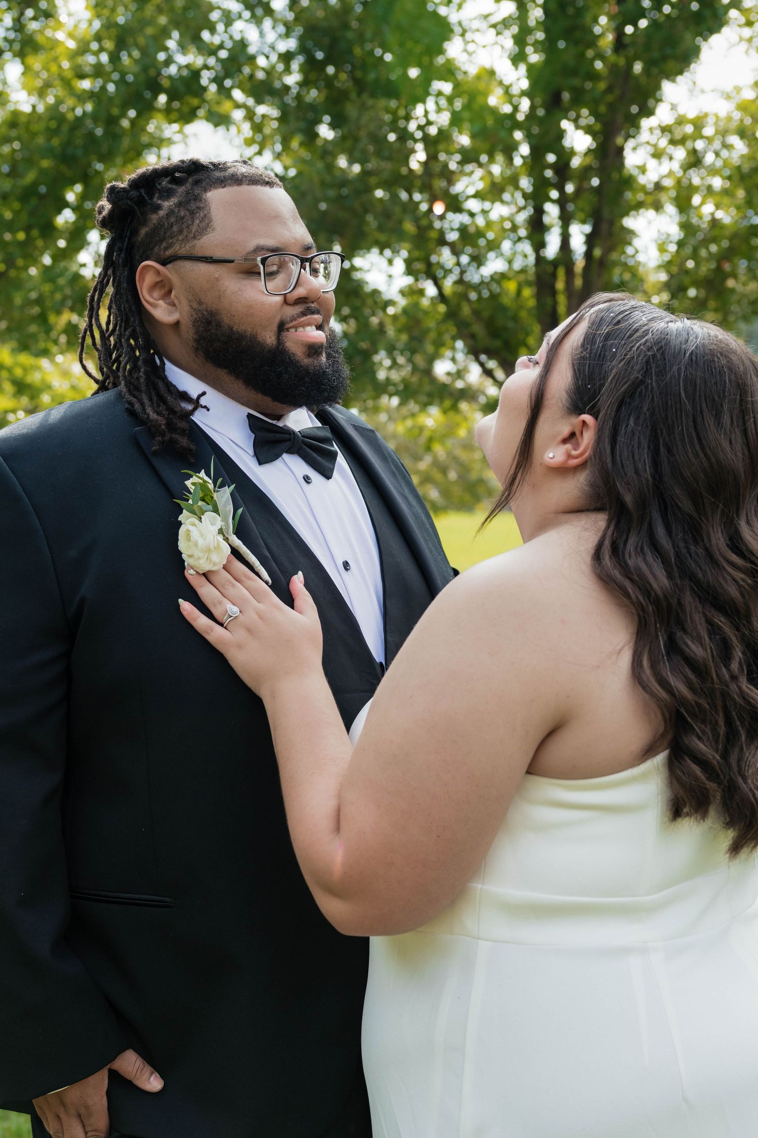 Bride gazes up at groom in tuxedo with boutonniere in romantic outdoor wedding portrait.