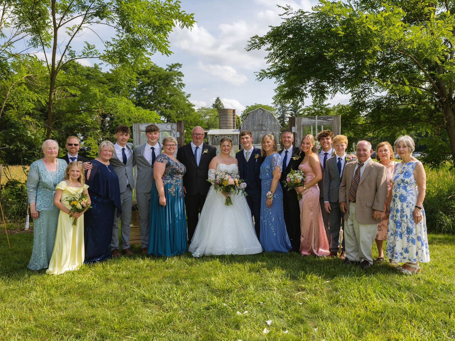 Wedding party poses outdoors on grass, bride in white gown with bouquet, guests in formal attire, surrounded by lush green trees.