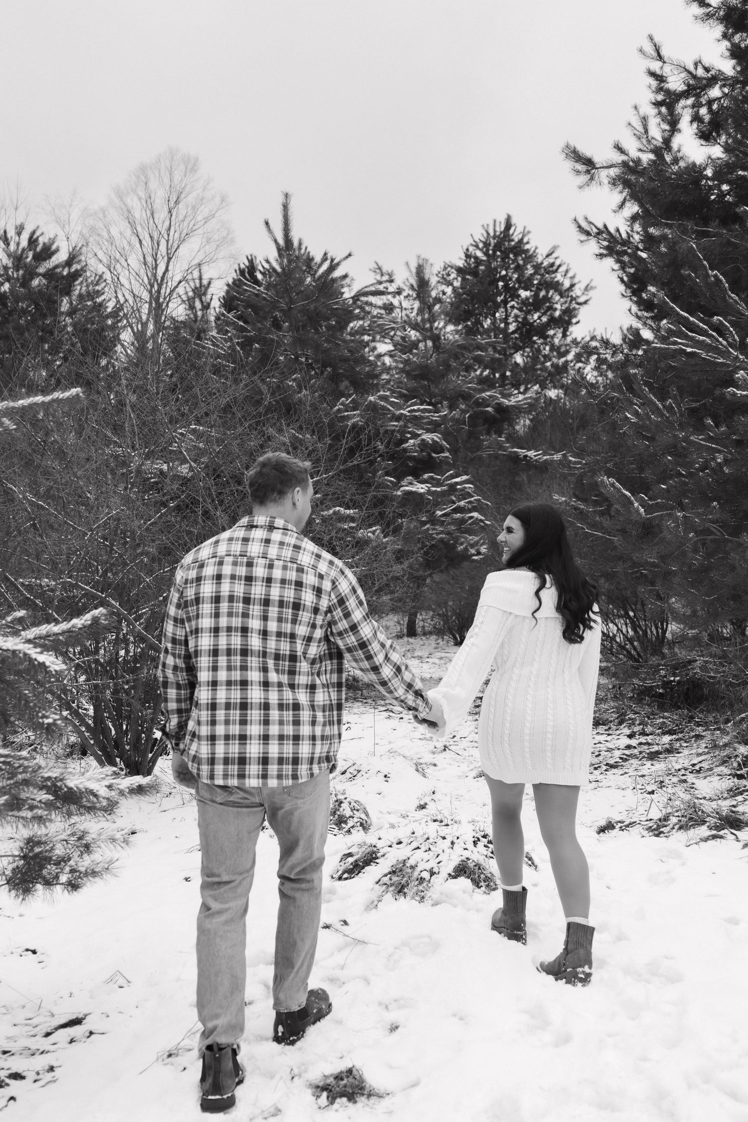 Black and white photo of a couple holding hands walking through a snowy pine tree forest together.