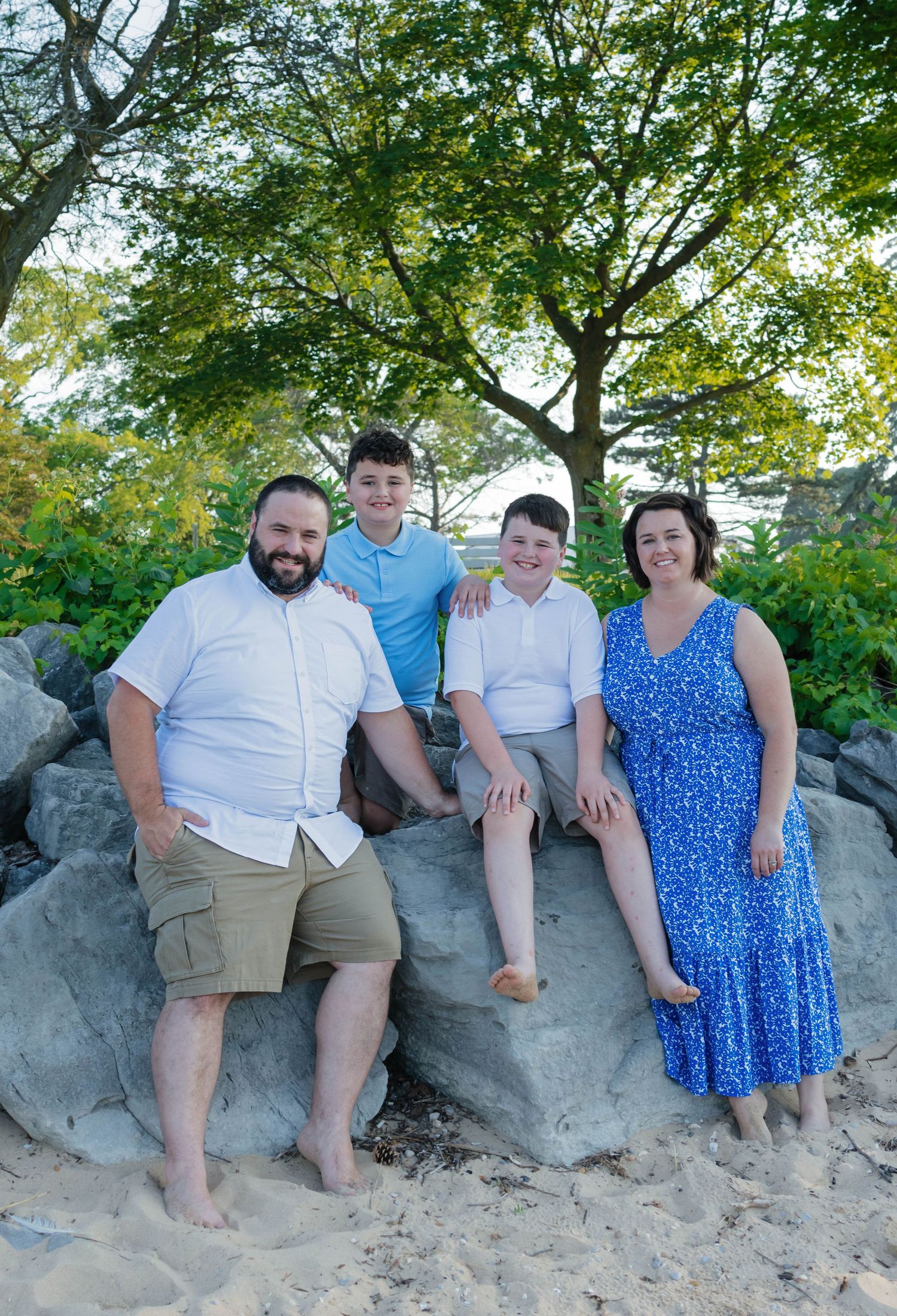 Family of four posing on large rocks with lush green trees on a sandy beach.