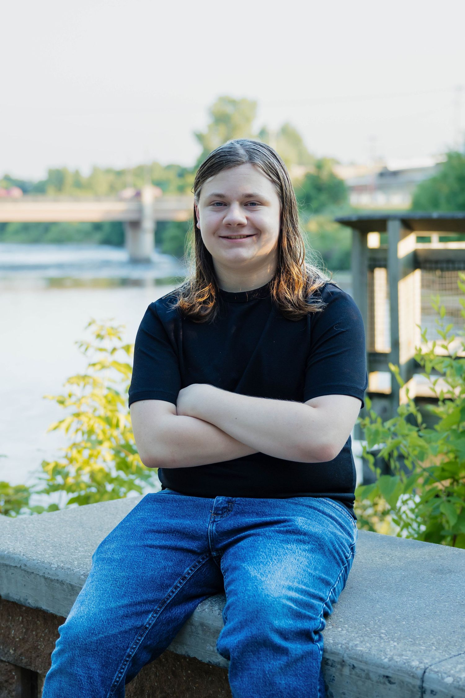 Teenage boy with long hair sitting on a stone wall with arms crossed, smiling near a river.