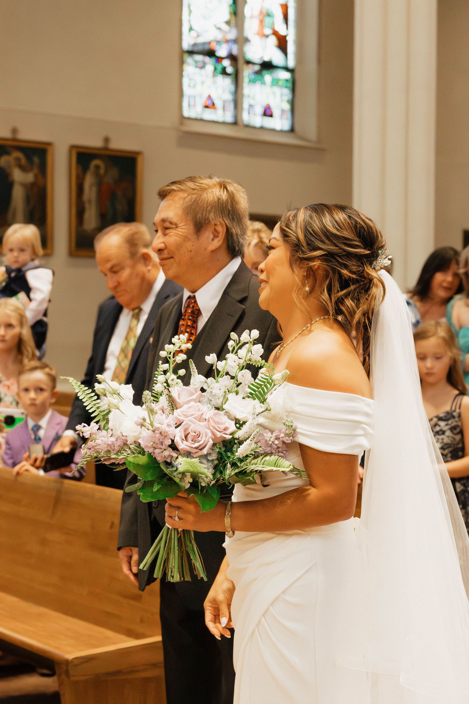 Bride in white off-shoulder gown holds pink bouquet while walking down church aisle with her father.