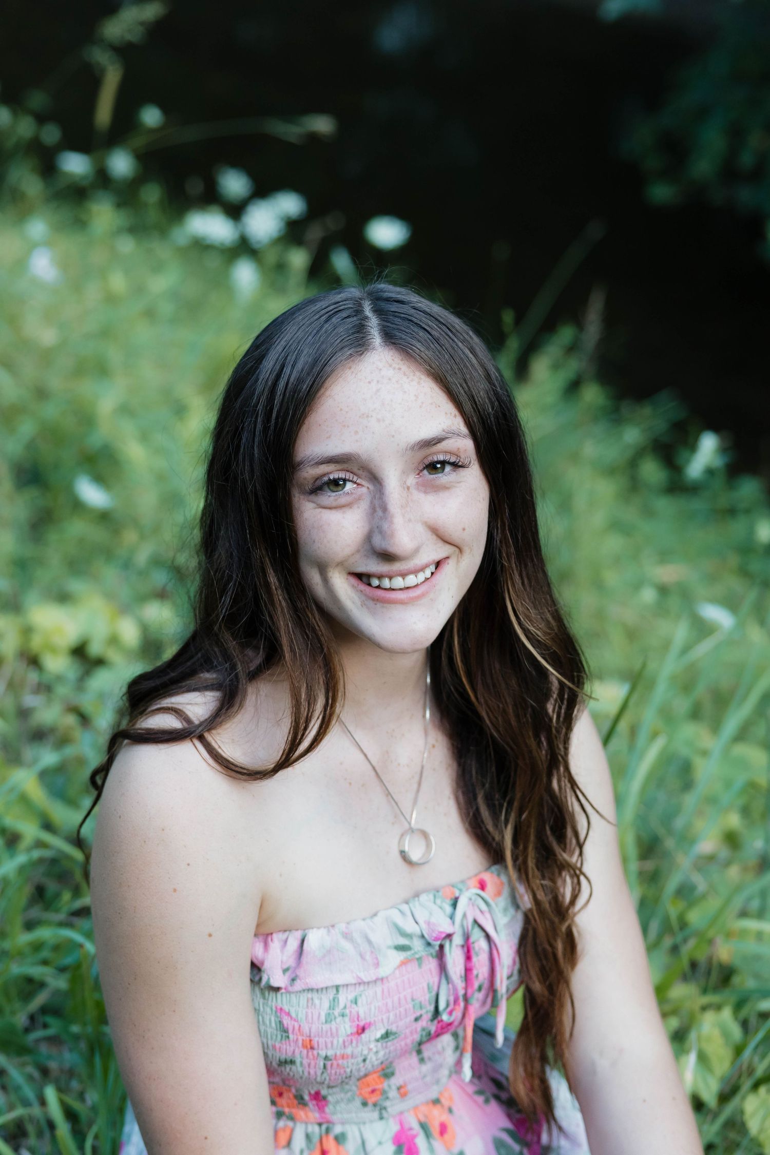 Smiling teenage girl with freckles sits in a garden wearing a floral strapless dress.