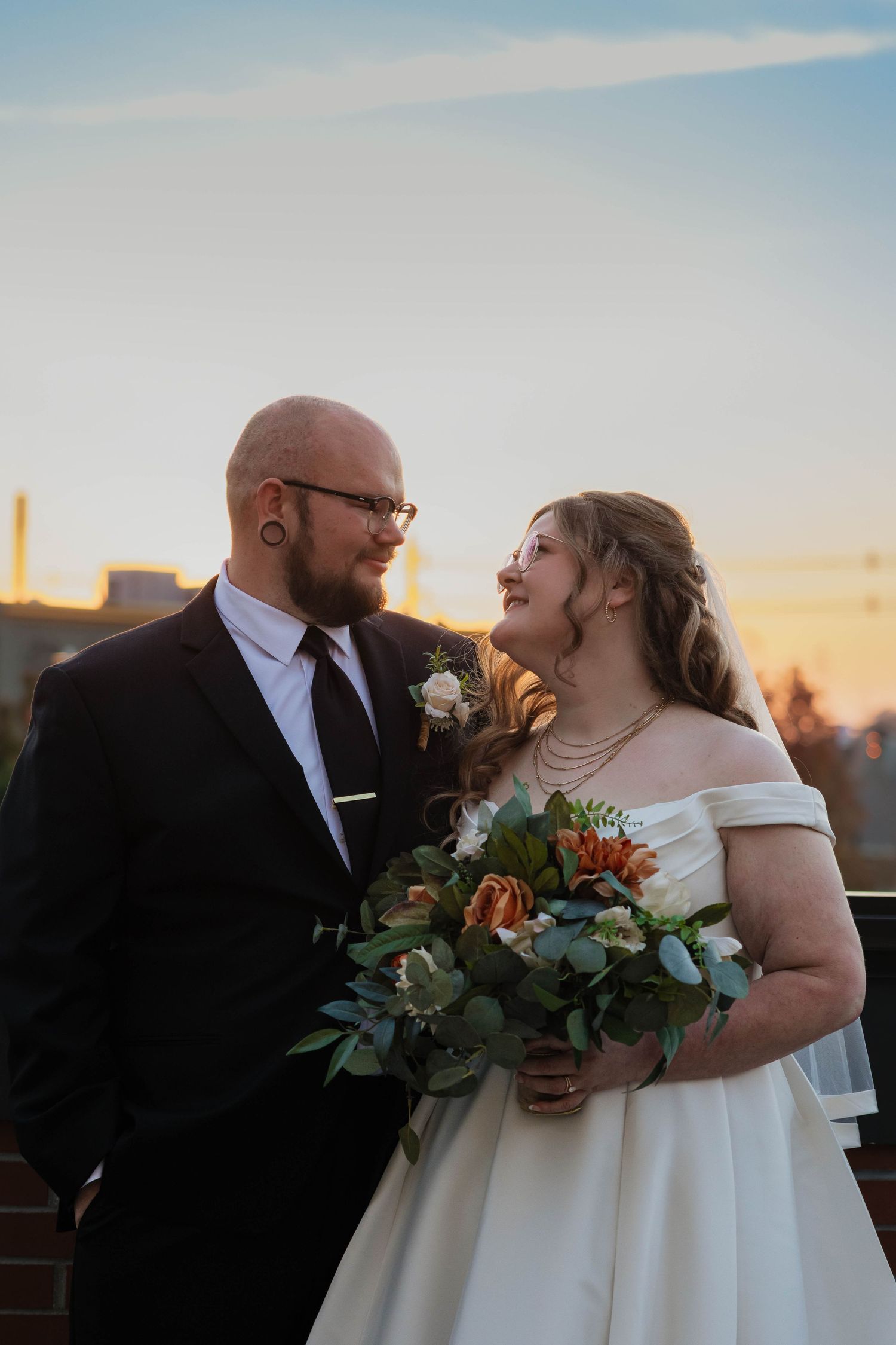 Bride and groom sharing a romantic moment at sunset, bride holding autumn bouquet in off-shoulder gown.