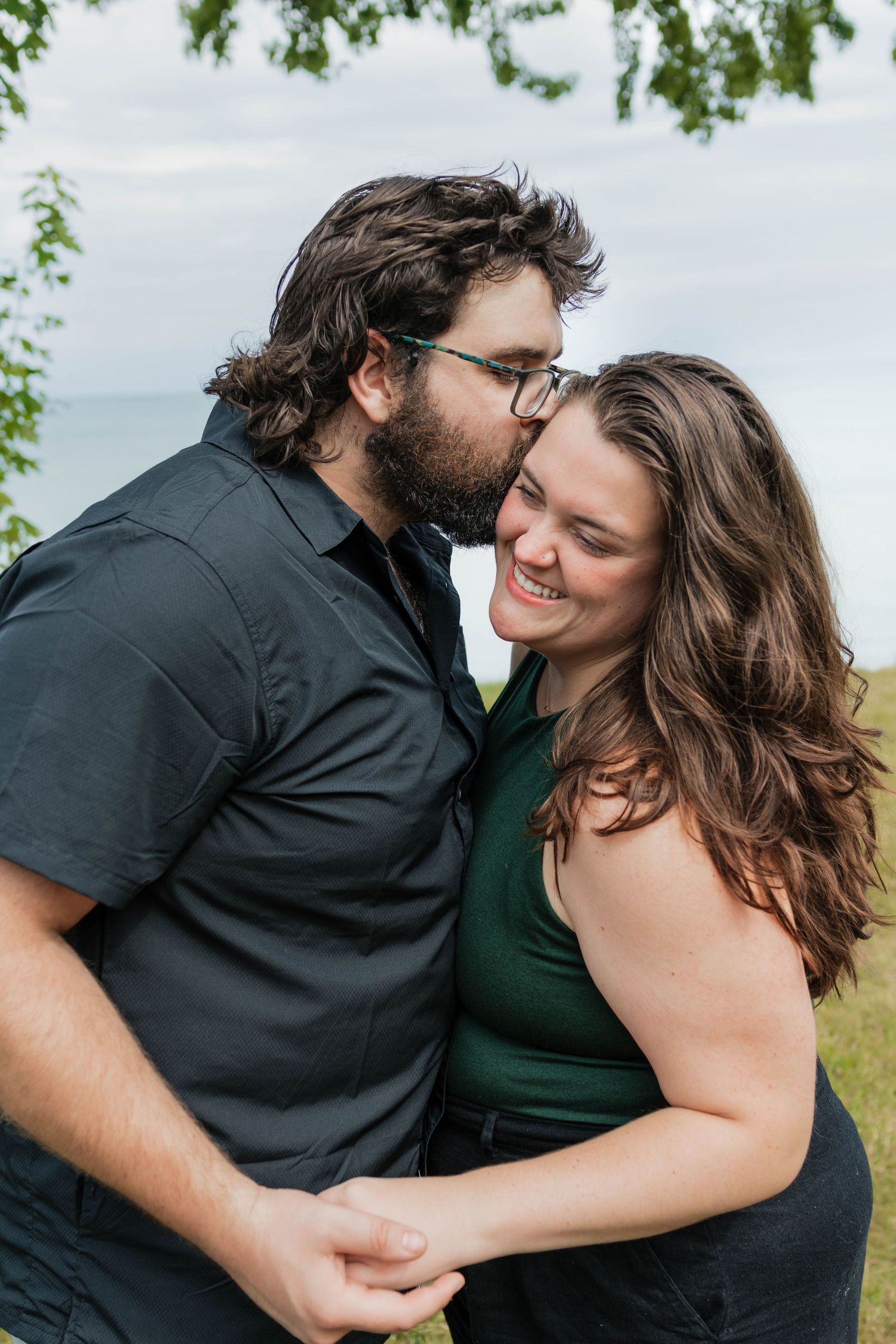 Bearded man kissing smiling woman on the cheek during romantic outdoor engagement session near the water.