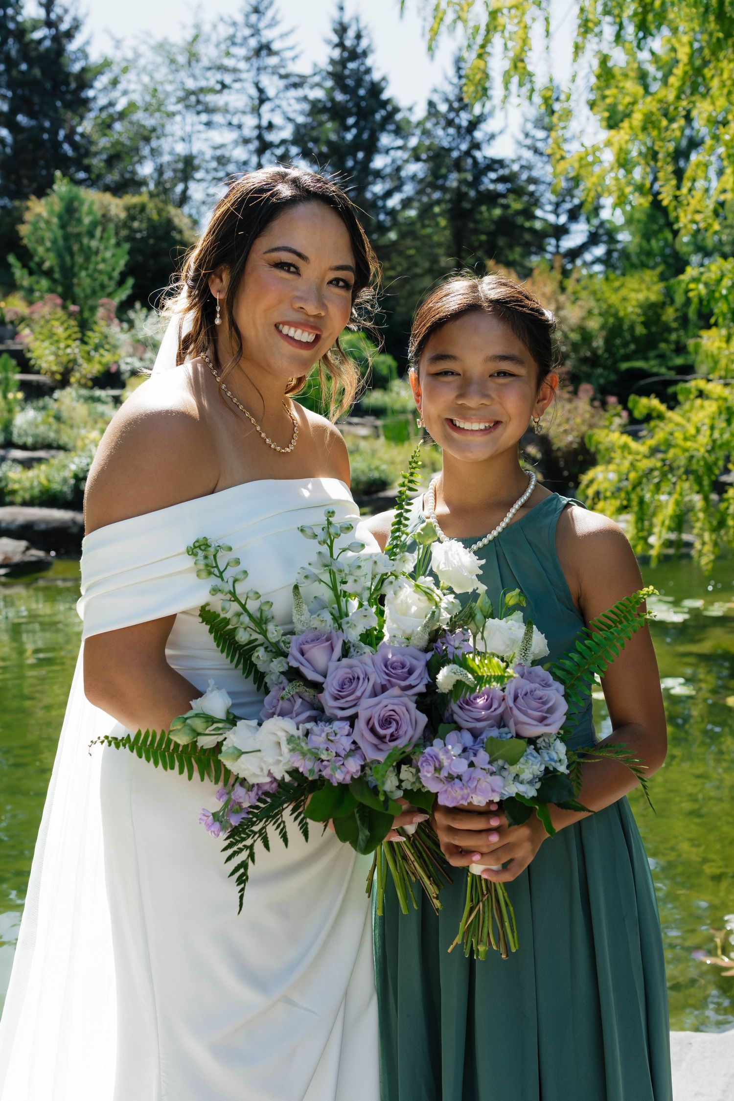 Smiling bride in white off-shoulder gown holds purple bouquet with young bridesmaid in teal dress.
