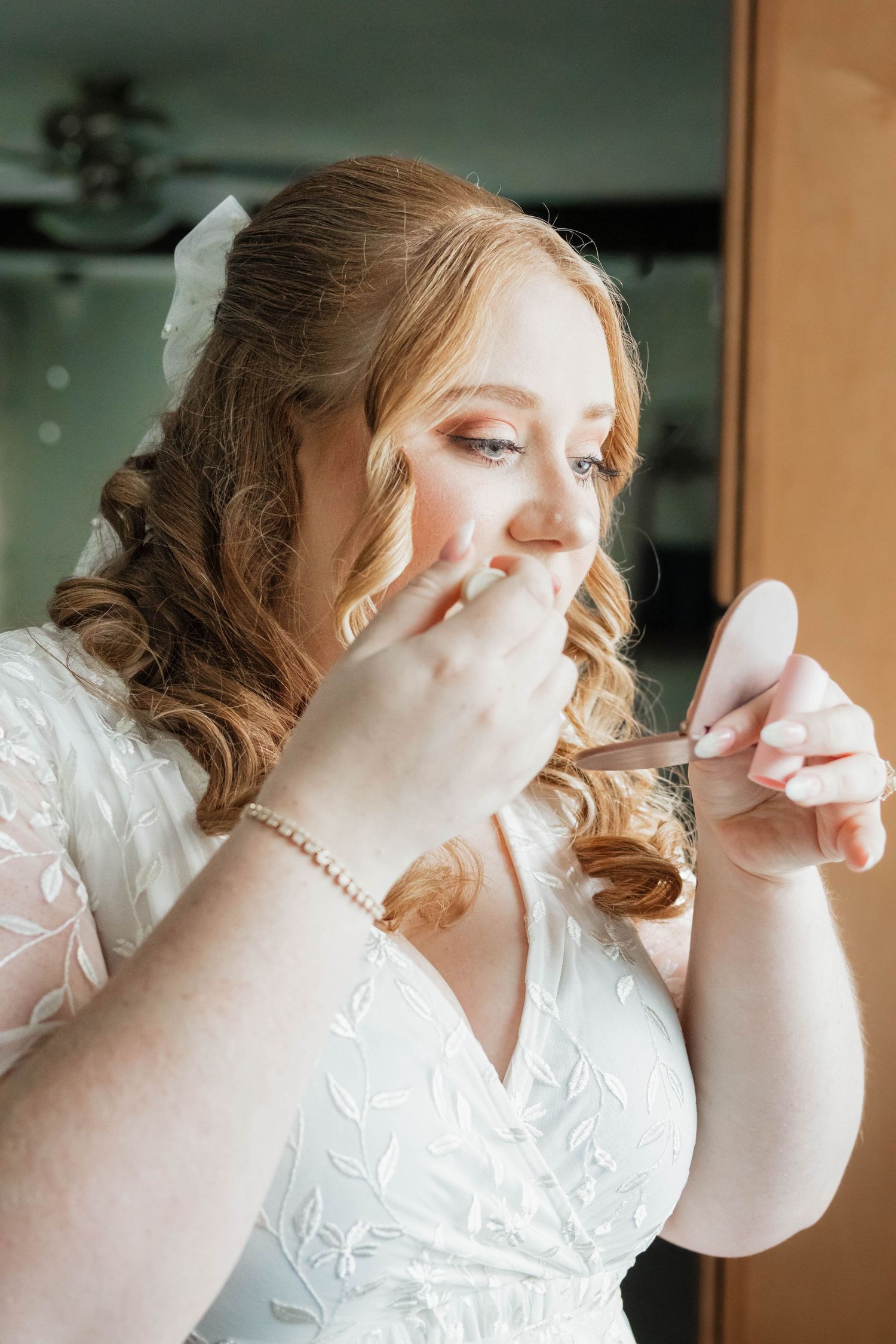 Bride in white lace gown touches up her lipstick using a small compact mirror.